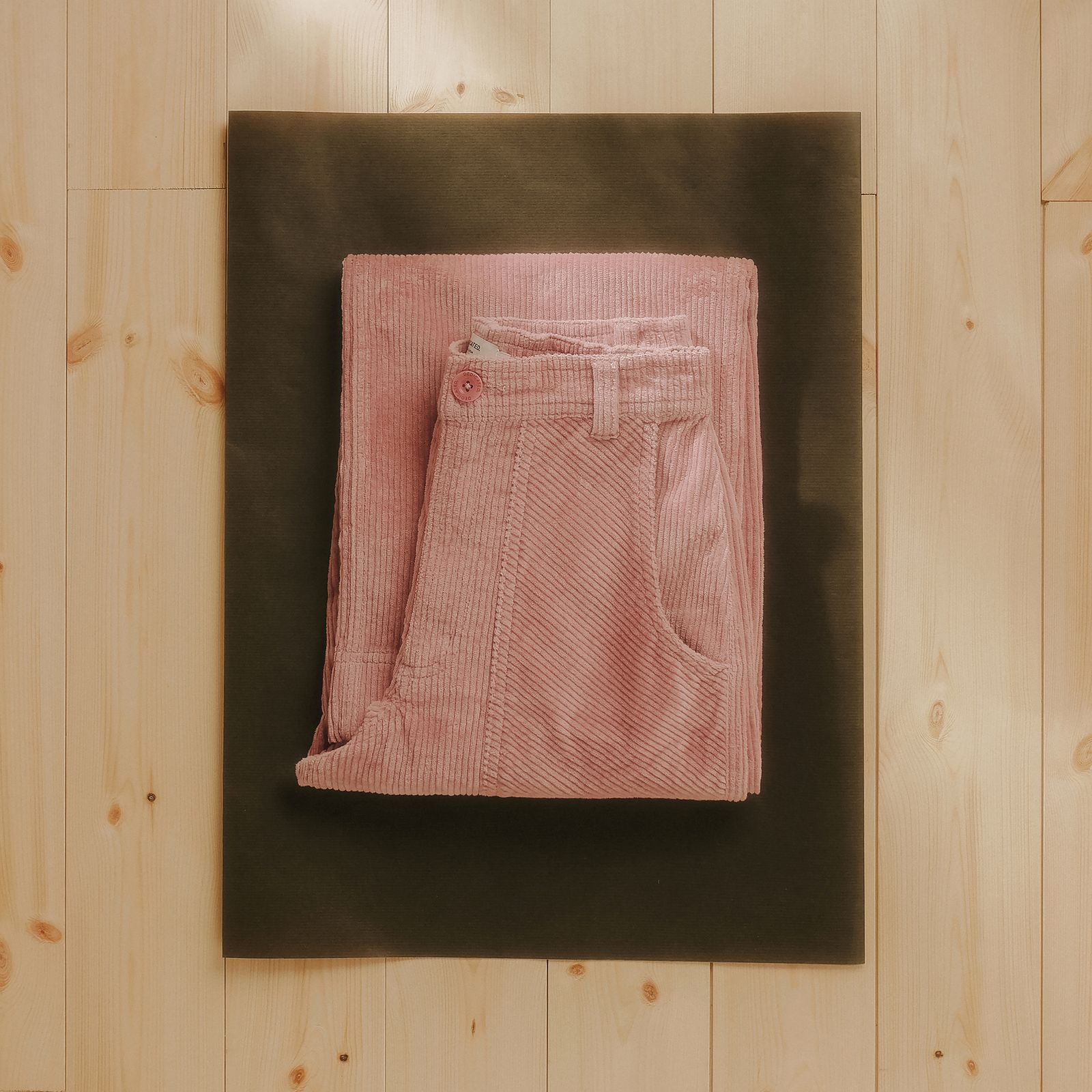 Folded pink corduroy pants on a dark mat, placed on a light wooden floor.