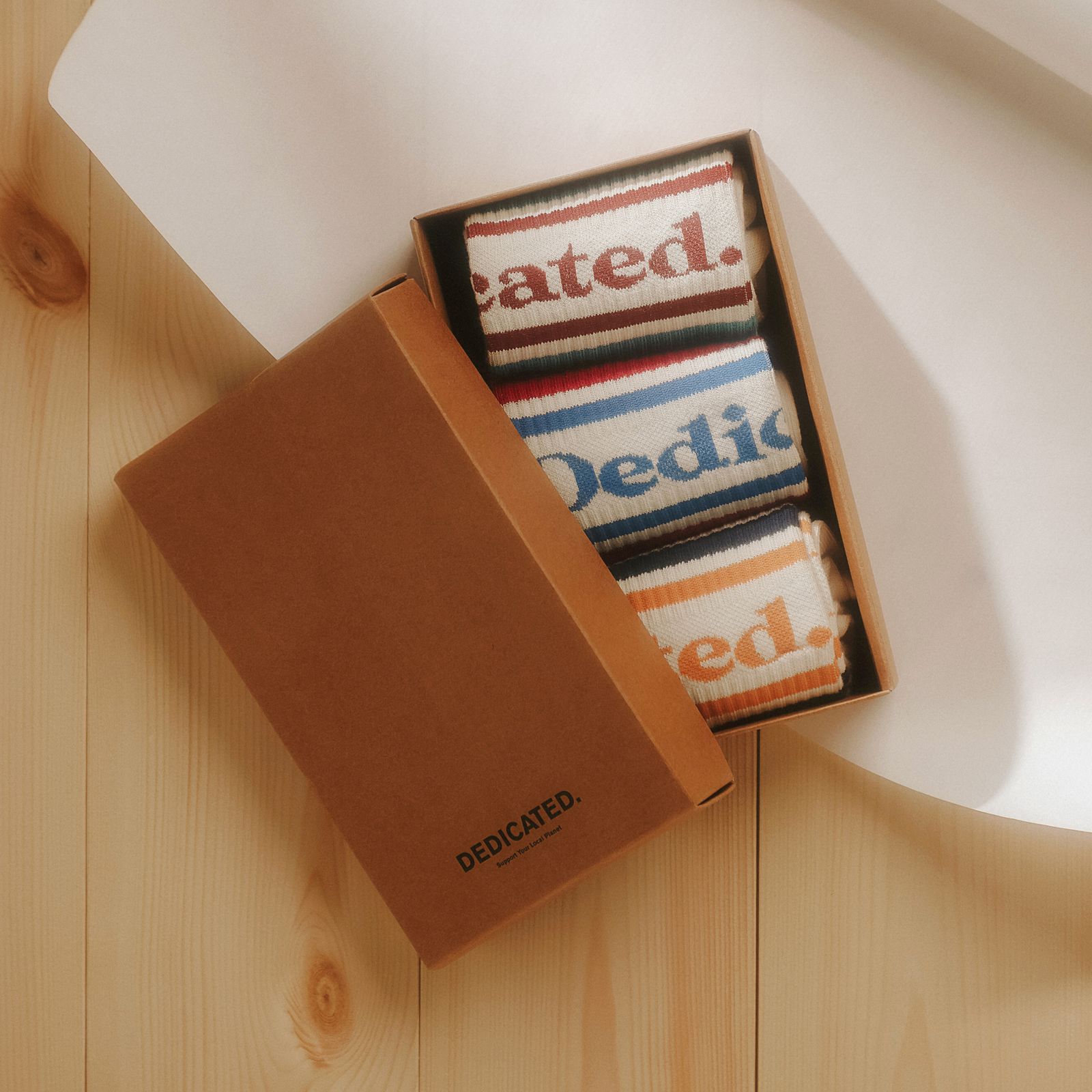 Open box of colorful socks with "Dedicated" text, placed on a light wooden surface.