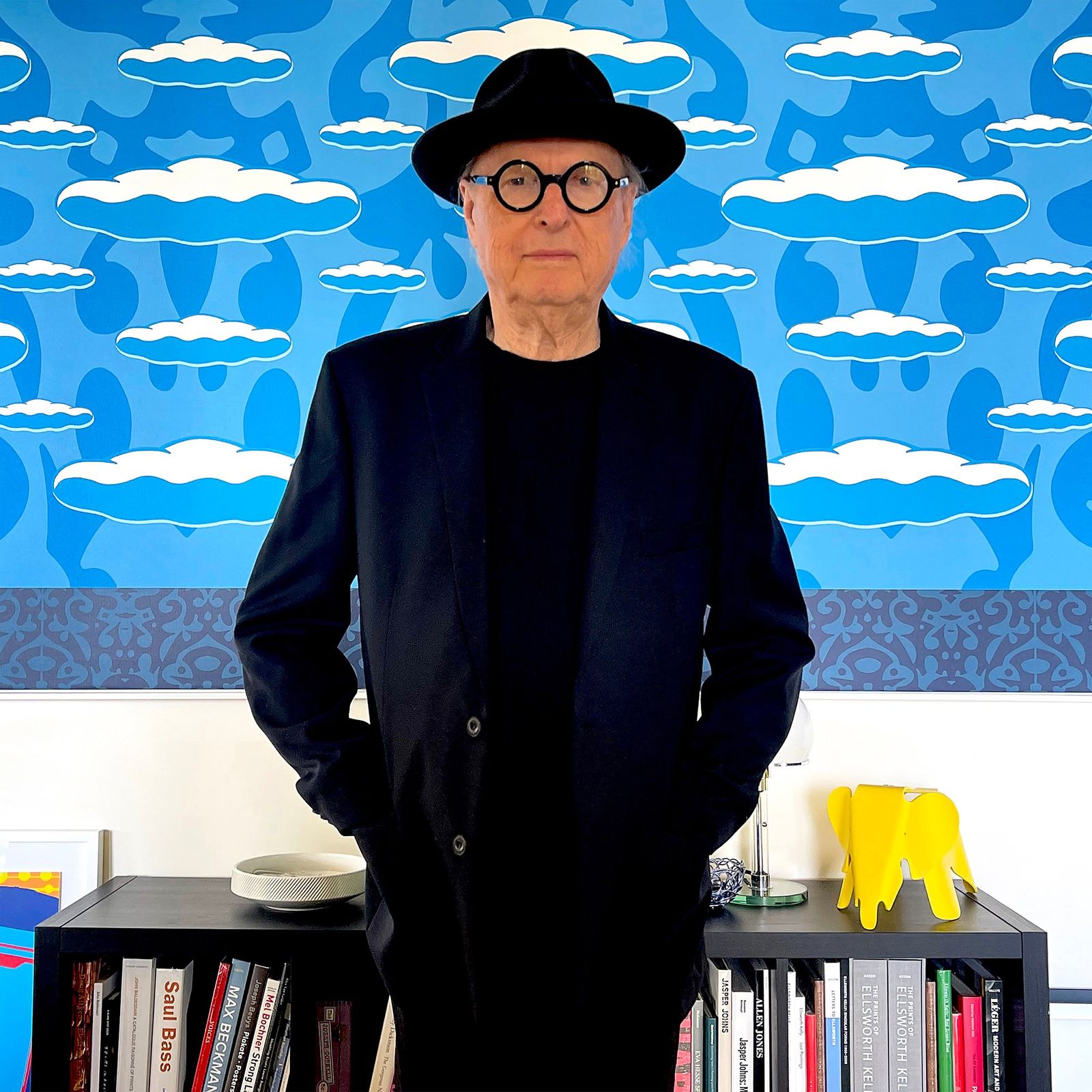 Man in black suit and hat stands in front of a blue cloud-patterned wall, with books and a yellow elephant figurine on a shelf behind him.