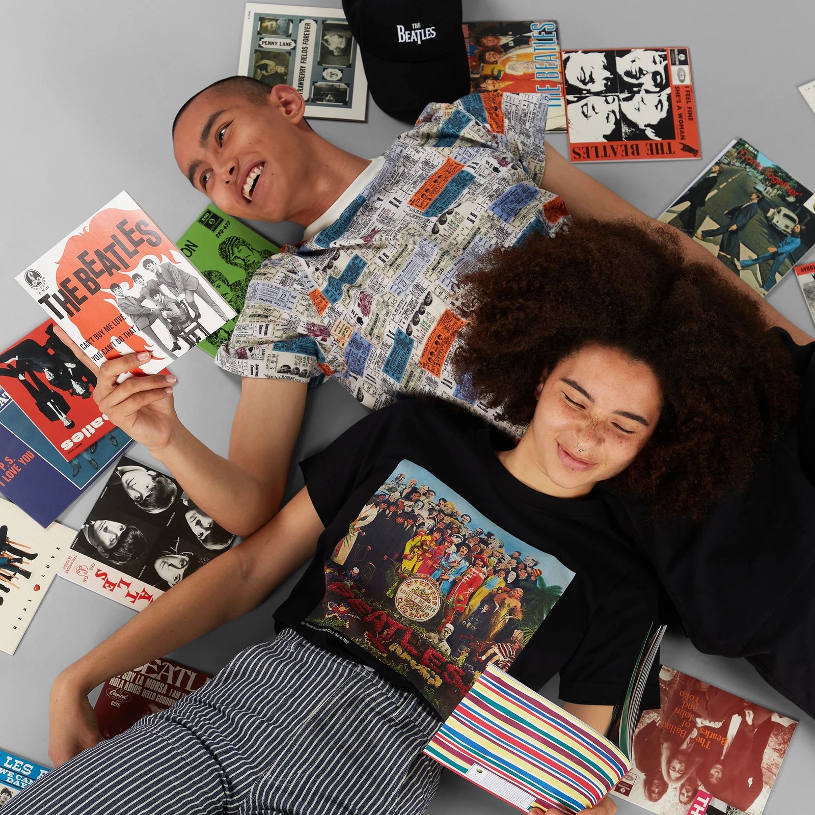 Two people lying on the floor surrounded by Beatles memorabilia, smiling and holding albums. Both wear Beatles-themed clothing.