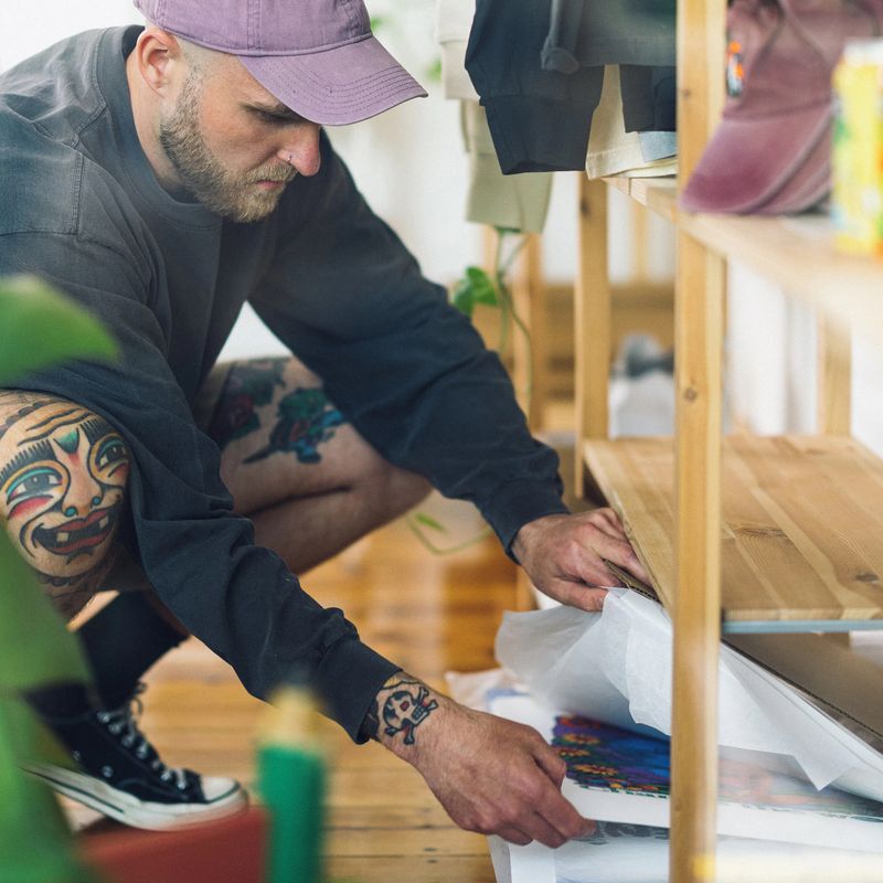 Man with tattoos and a cap crouches, organizing colorful artwork on a wooden shelf in a bright room.