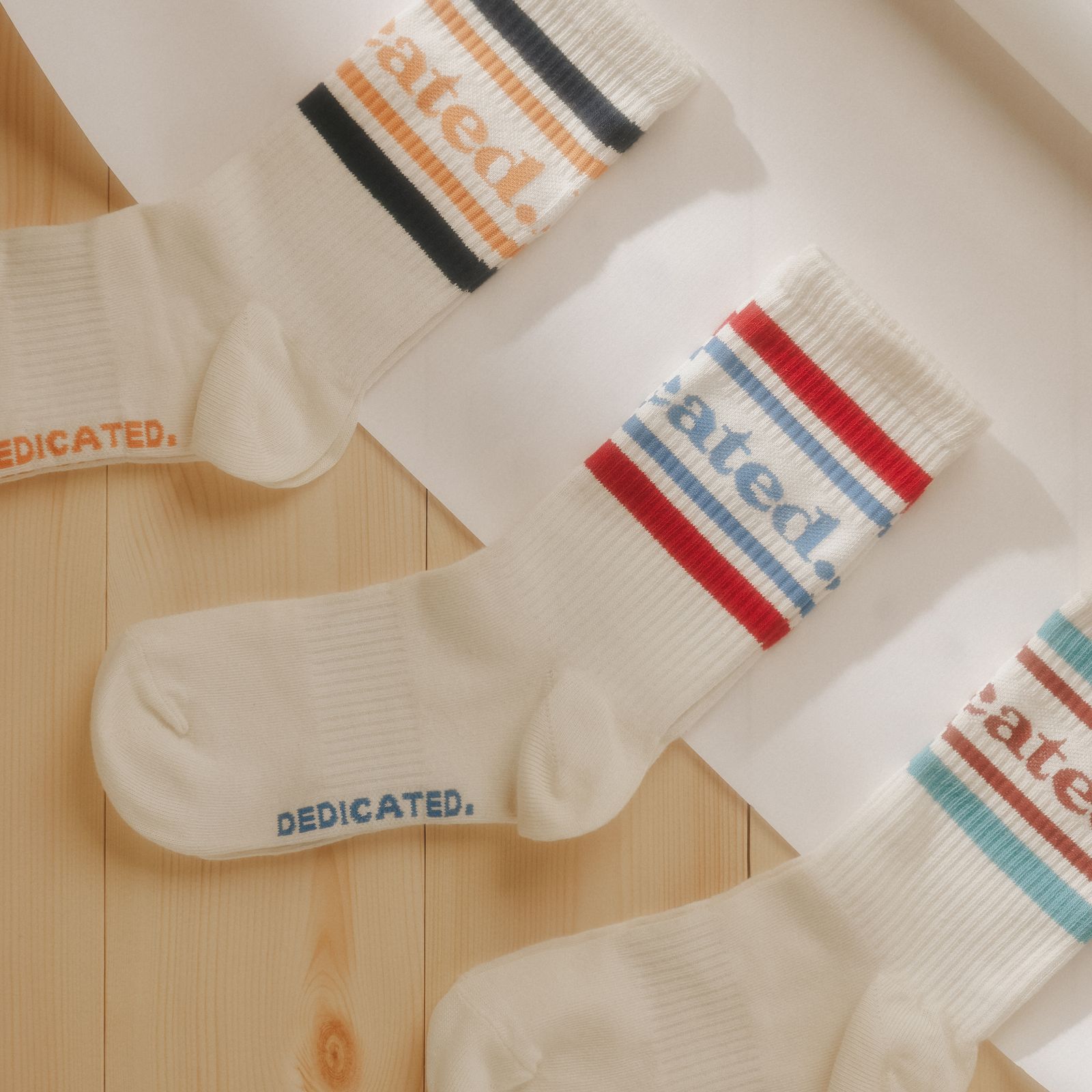 Three pairs of white socks with colorful stripes and the word "DEDICATED" on a wooden surface.