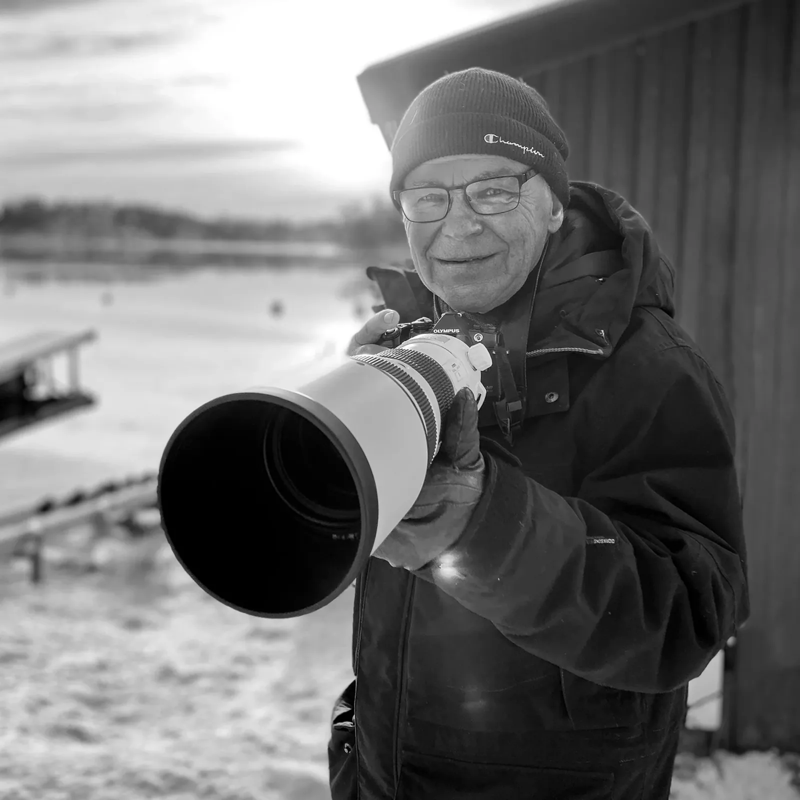 Person in winter clothing smiles while holding a large camera lens outdoors, with a snowy landscape and wooden building in the background.