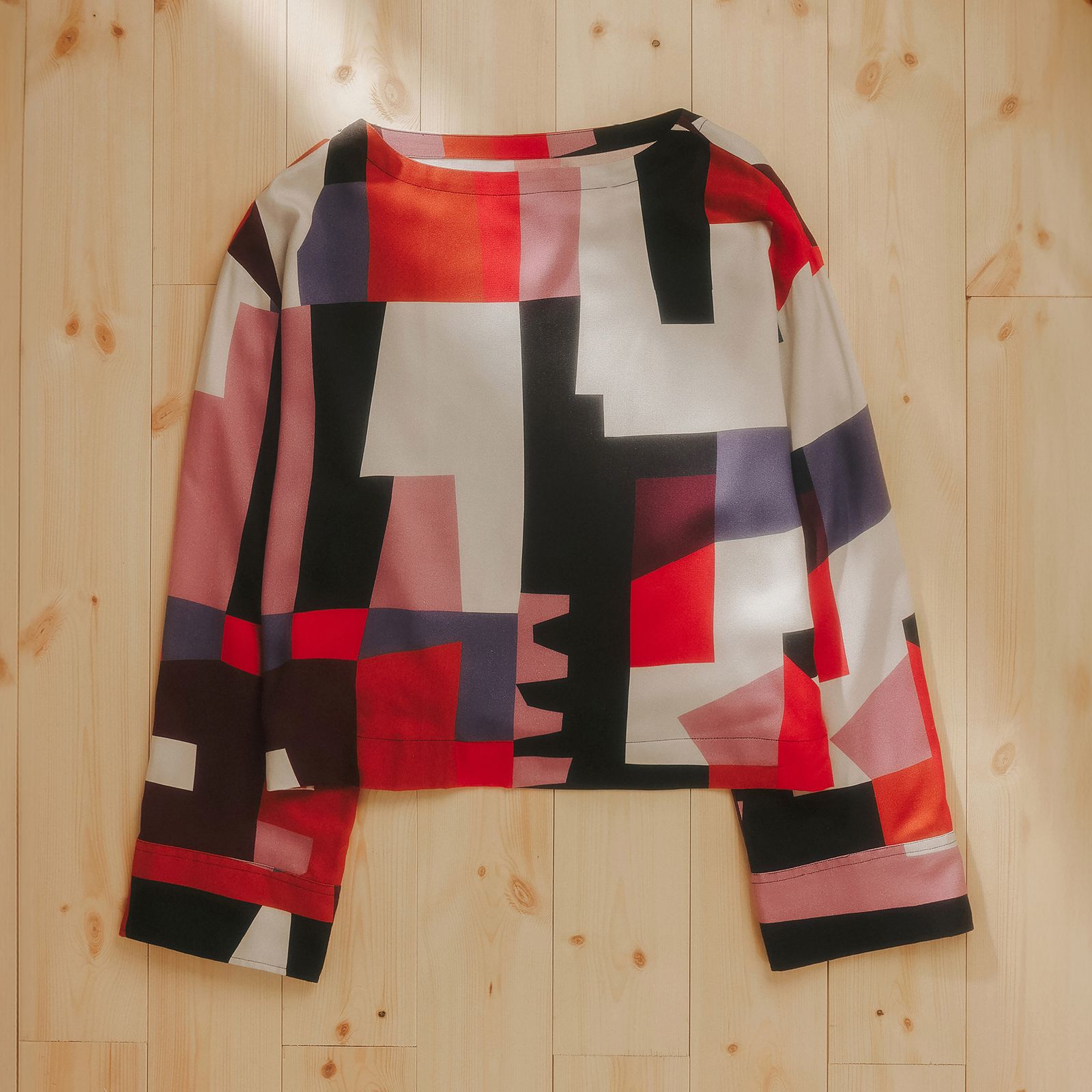 Colorful geometric-patterned long-sleeve shirt with red, black, and white blocks, laid flat on a light wooden floor.