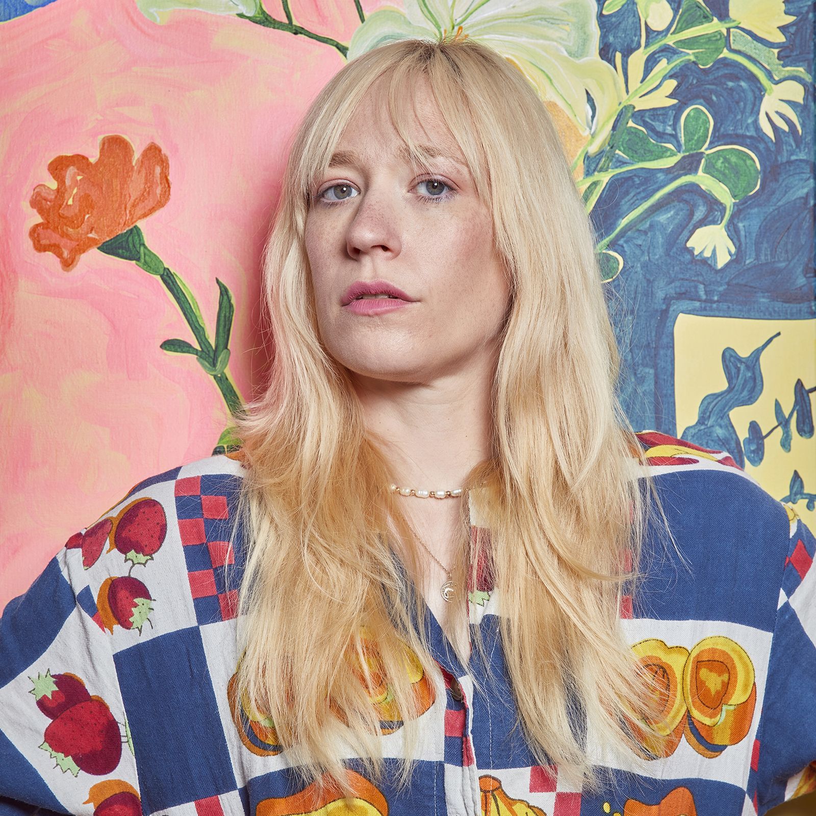 Person with long blonde hair wearing a colorful shirt featuring fruits, standing in front of a vibrant floral mural.