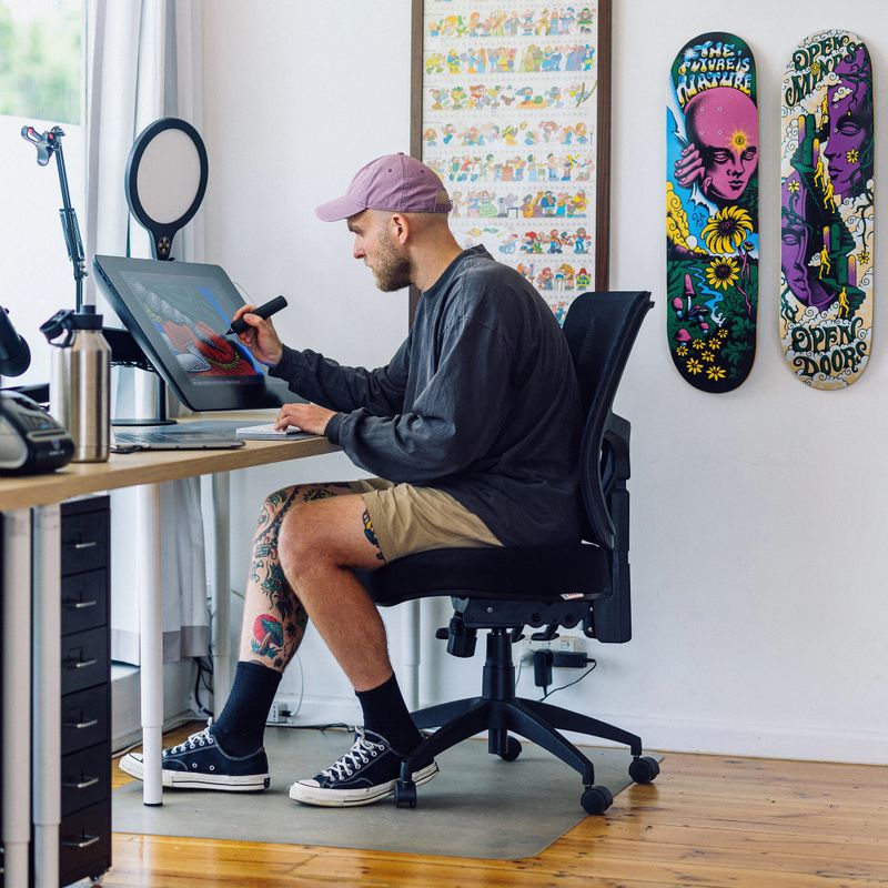 A man in casual attire works on a digital drawing tablet in a creative studio, surrounded by colorful skateboard art on the wall.