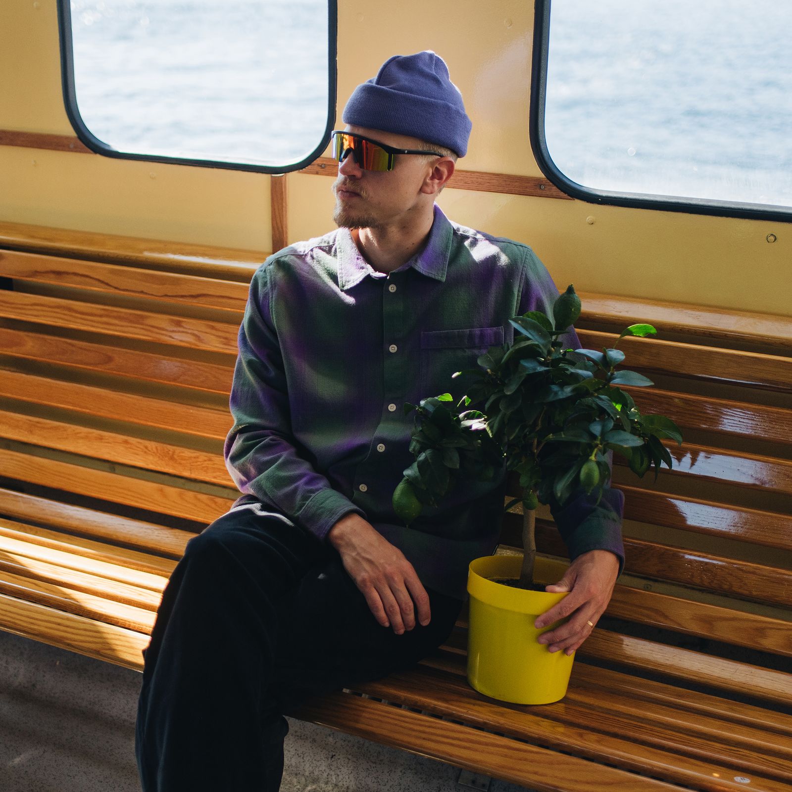 Man in a purple beanie and sunglasses sits on a boat bench holding a potted plant, with water visible through the window.