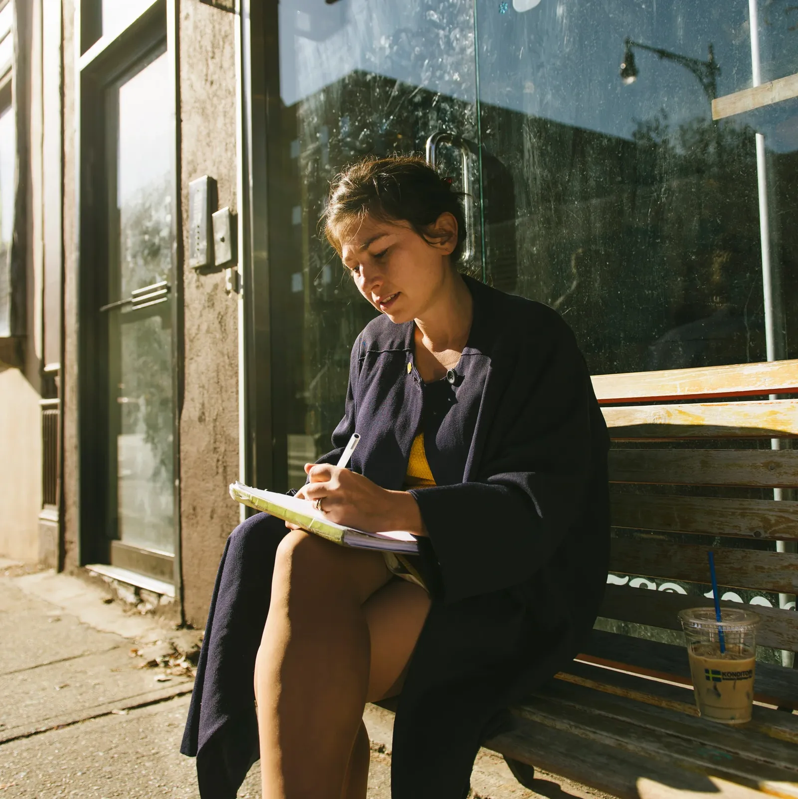 A woman sits on a bench outside, writing in a notebook. She wears a dark coat and is illuminated by sunlight. An iced drink is beside her.