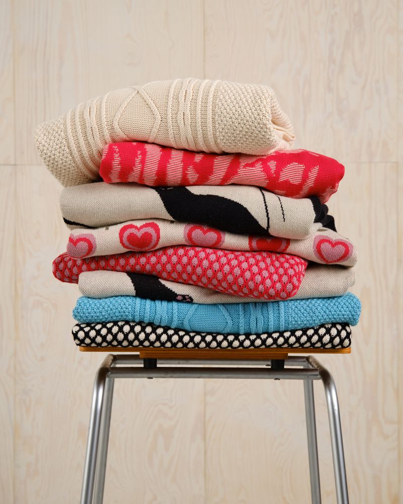 A stack of colorful folded sweaters with various patterns, including hearts and polka dots, sits on a wooden stool with metal legs.