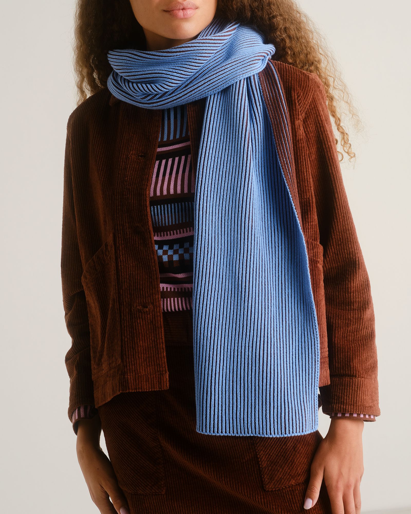 A person wearing a blue ribbed scarf over a patterned sweater and brown corduroy jacket and skirt, with curly hair visible.