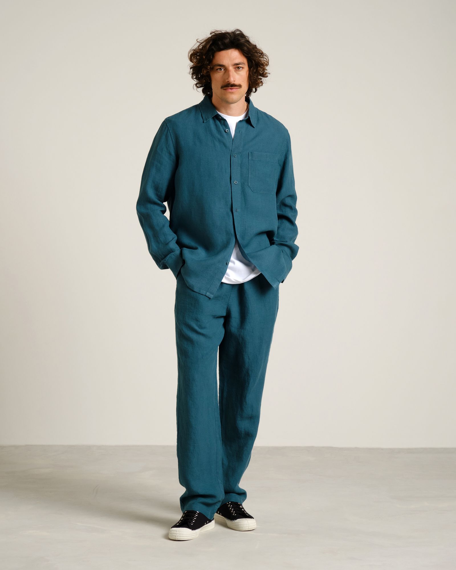 Man with curly hair and mustache wearing a teal shirt and pants, a white undershirt, and black sneakers, standing with hands in pockets.