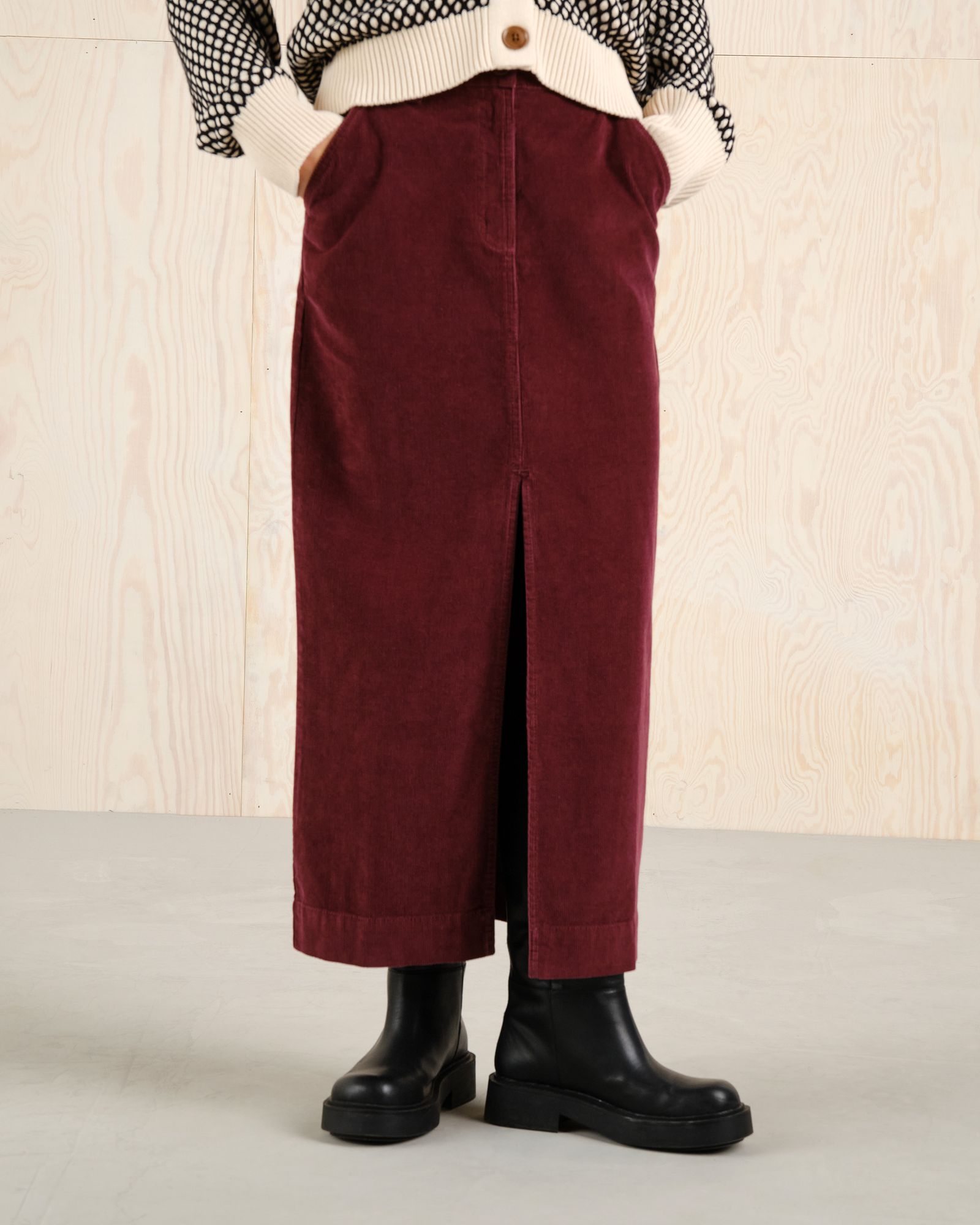 Person wearing a long burgundy corduroy skirt, black boots, a white sweater, and a patterned shirt, standing on a light-colored floor.