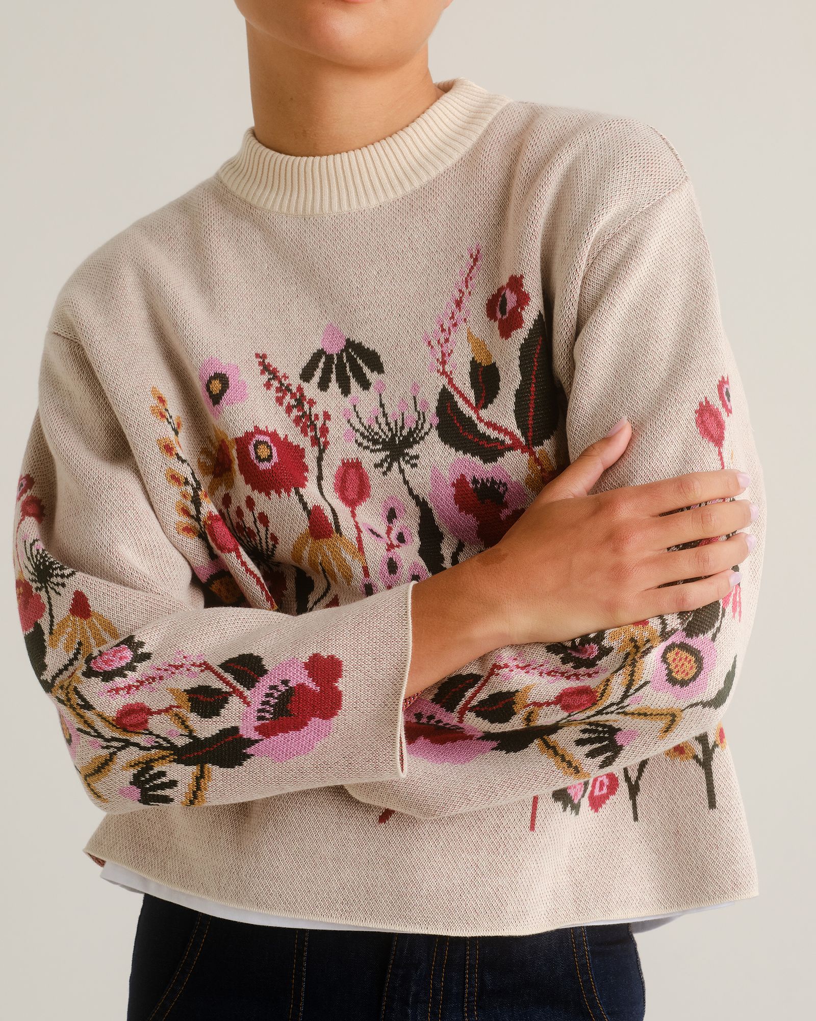 Person wearing a cream sweater with colorful floral embroidery and blue jeans, arms crossed.