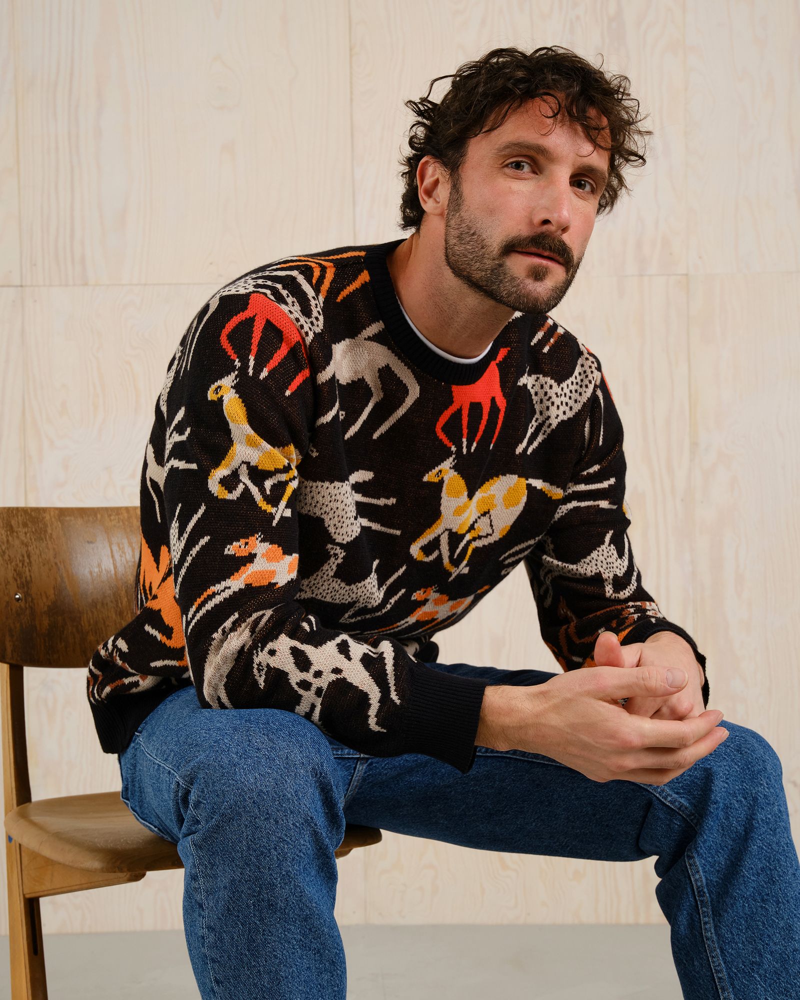 Man with curly hair and beard sits on a wooden chair, wearing a patterned sweater with animal motifs and blue jeans, against a light wood background.