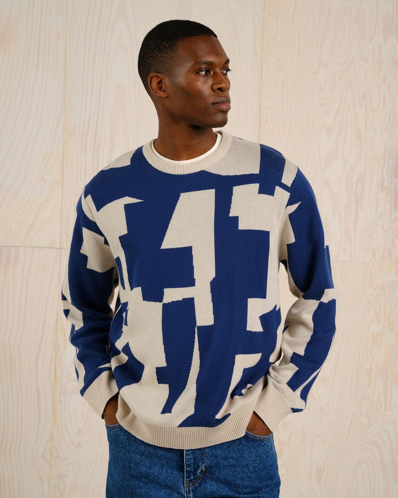 Man wearing a blue and beige geometric-patterned sweater and jeans, standing against a light wooden background, looking to the side.