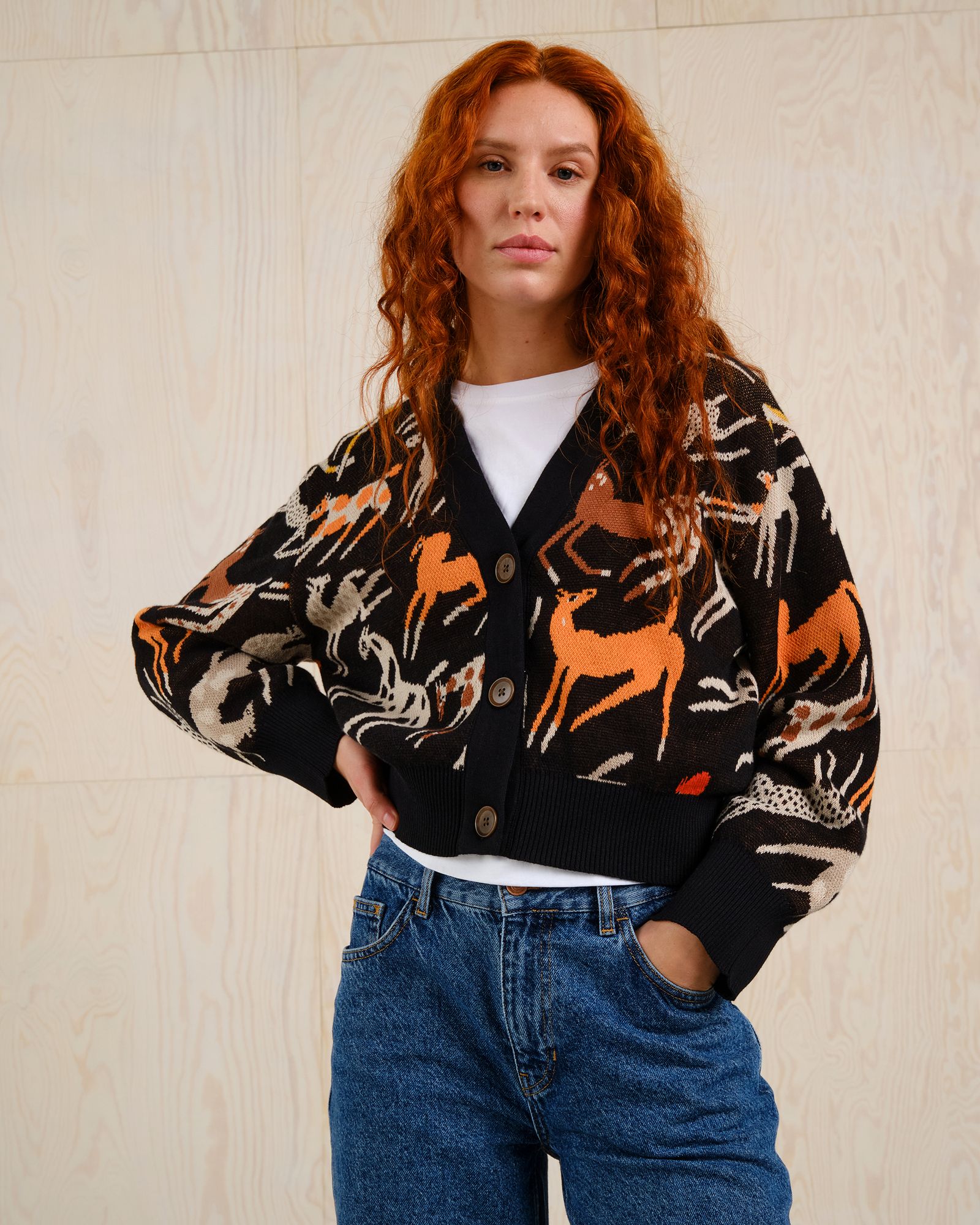 Person with long red hair wearing a black cardigan with animal patterns and blue jeans, standing against a light wood background.