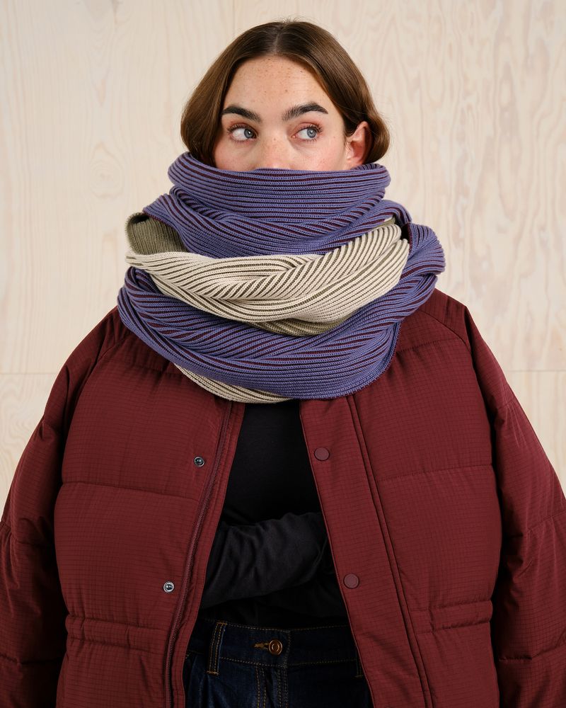 Person wearing a large, layered scarf in purple and beige, with a maroon quilted jacket, standing against a light wood background.