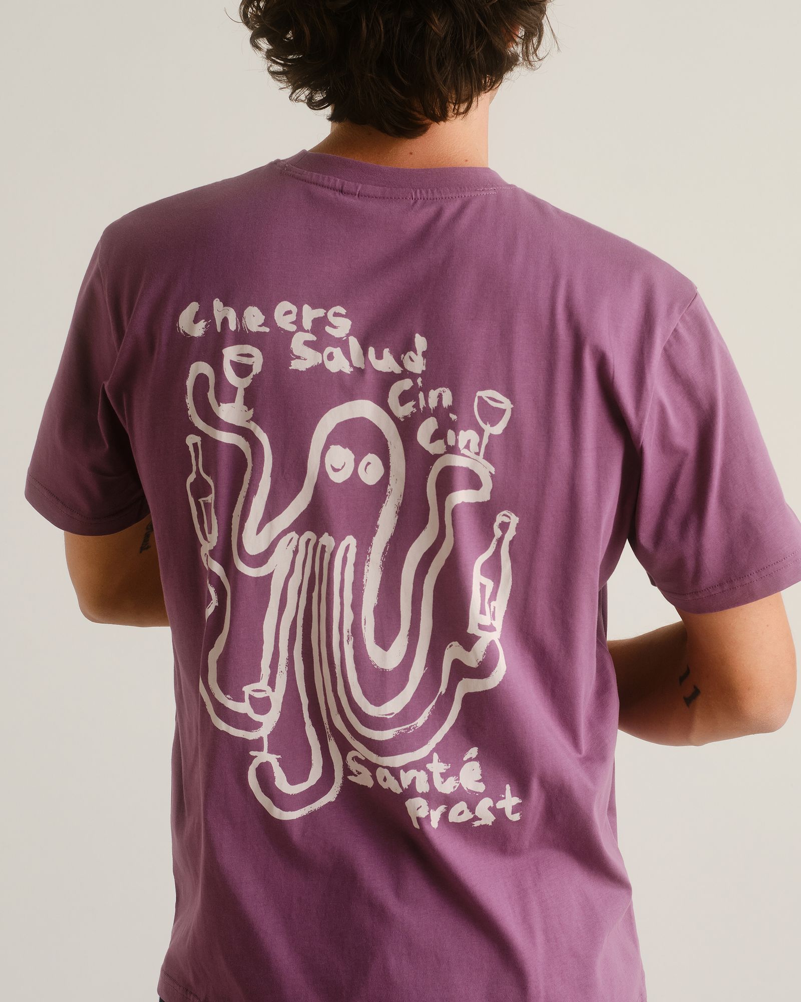 Person wearing a purple T-shirt with an octopus illustration holding wine glasses and bottles, surrounded by multilingual cheers phrases.