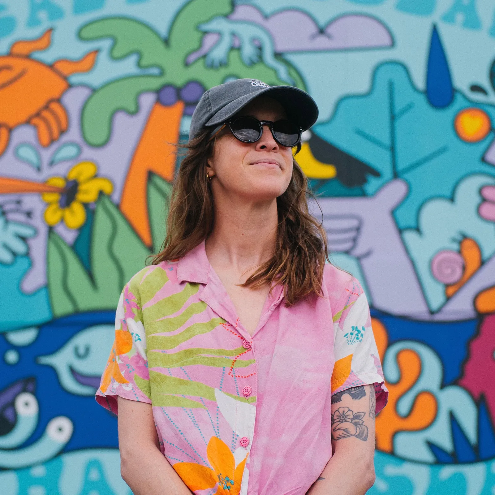 Woman in a colorful shirt and cap stands in front of a vibrant, cartoon-style mural featuring sea creatures and plants.