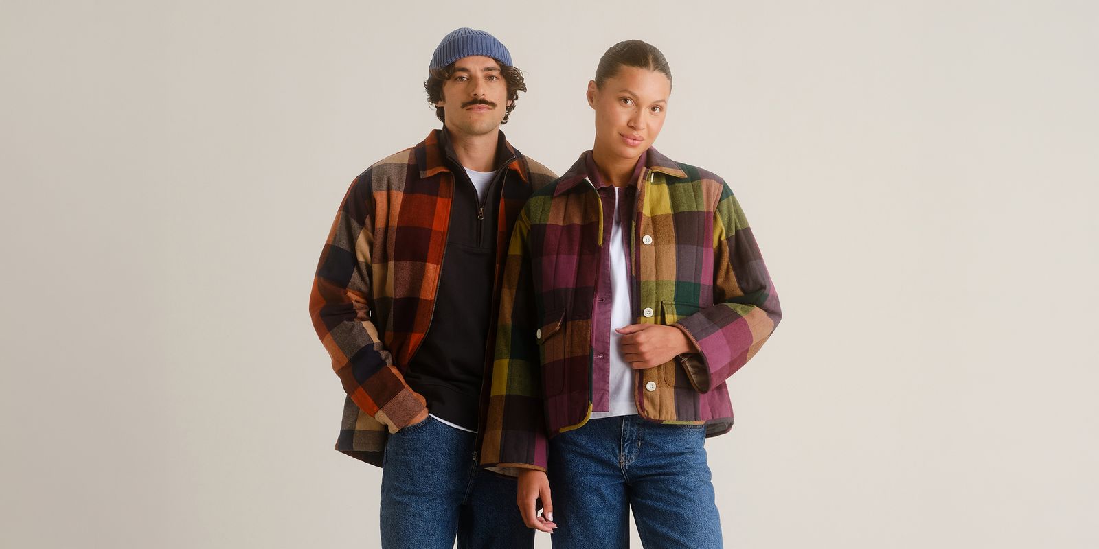 Two models wearing colorful plaid jackets and jeans stand against a light background. One wears a beanie, both have relaxed expressions.
