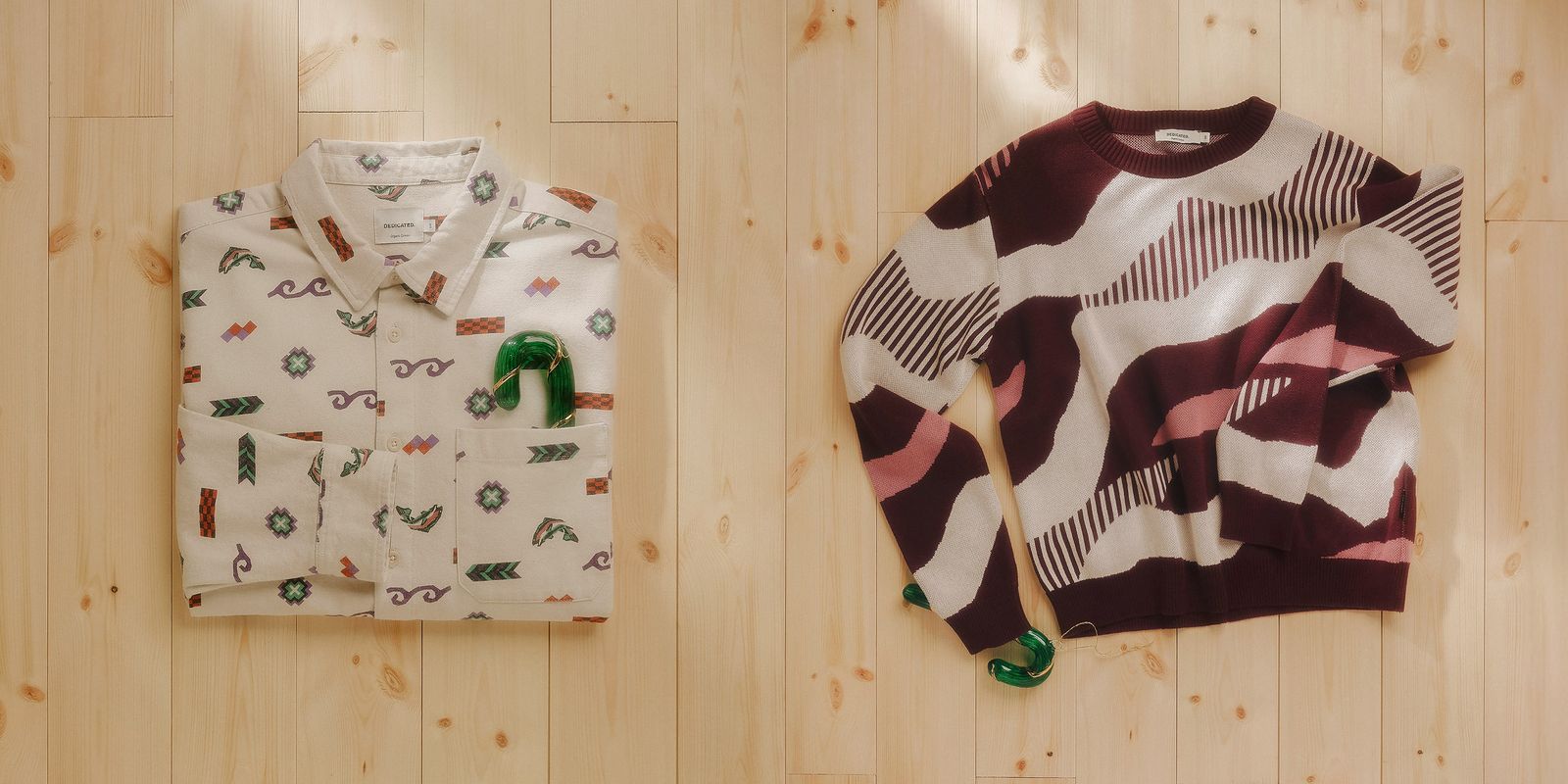 Folded white printed button-up shirt with green scrunchie and folded maroon-and-cream abstract patterned sweater on light wood floor.