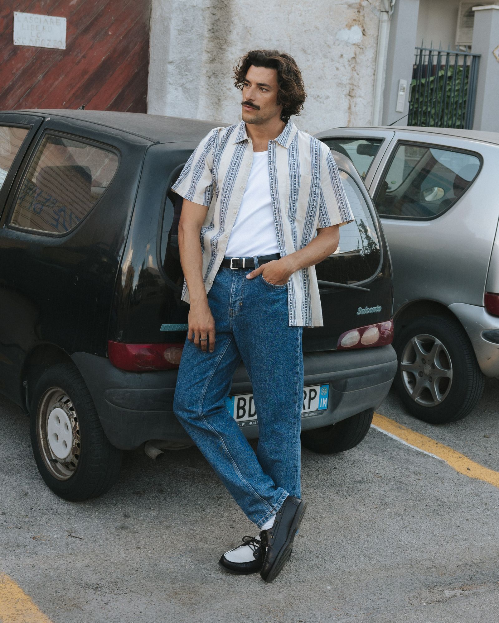 Man in a striped shirt and jeans leans against a parked car in a lot, exuding a relaxed, vintage style.