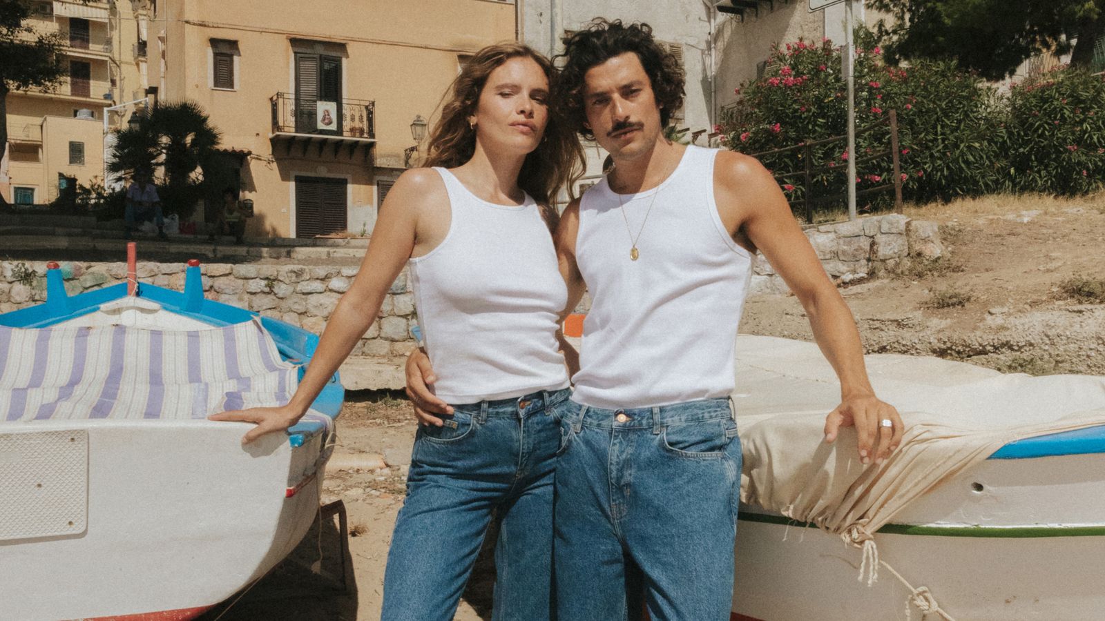 A man and woman in white tank tops and jeans stand between boats on a sandy beach, with buildings and greenery in the background.