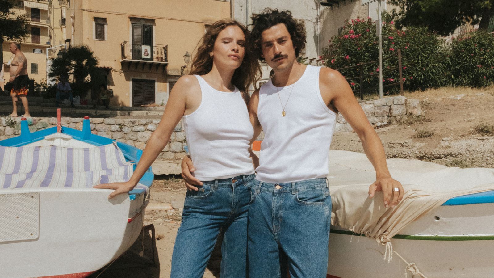 A man and a woman in white tank tops and jeans stand together on a beach, leaning against small, colorful boats, with buildings in the background.