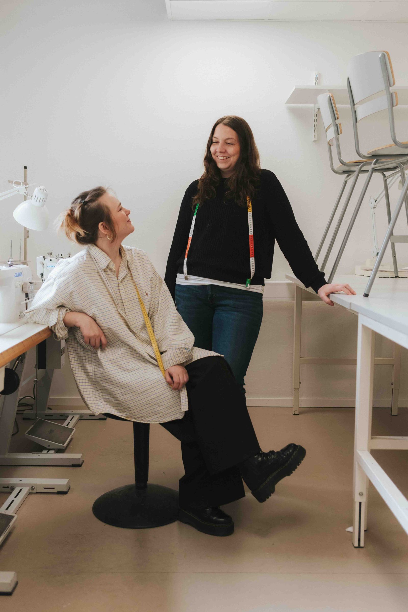 One girl sitting on a chair looking up to another girl who's standing behind her and smiling