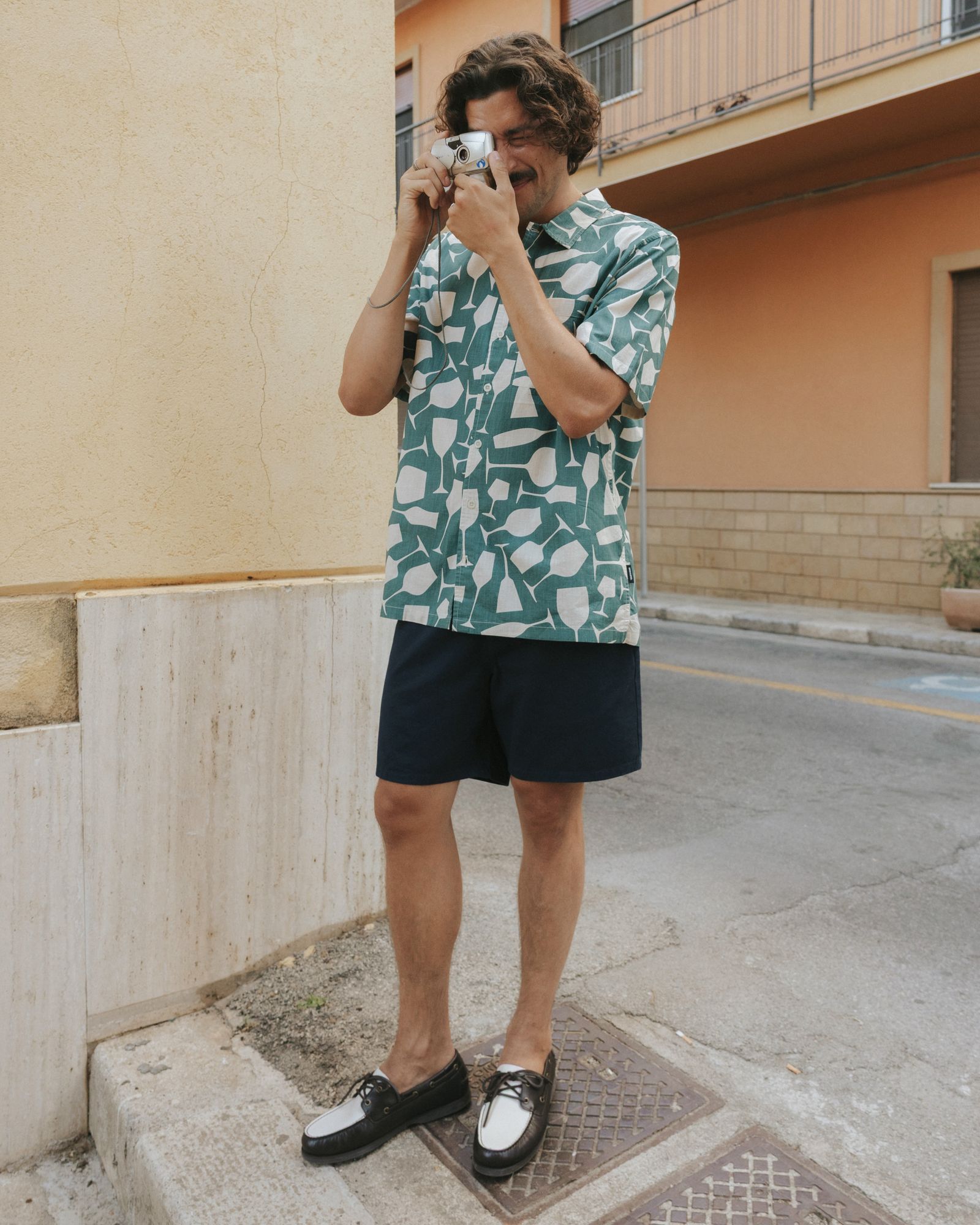 Man in patterned shirt and shorts taking a photo with a vintage camera on a street corner, standing near a beige wall.