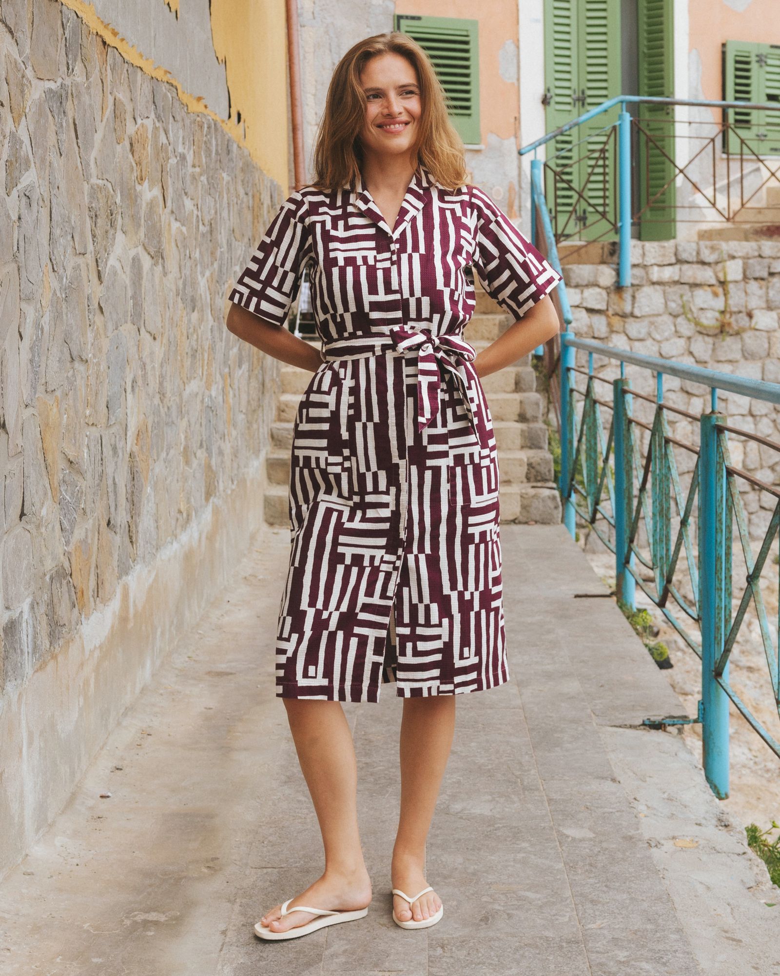 Woman in a geometric patterned dress, smiling, stands on a stone walkway by a railing with a stone wall and colorful building in the background.
