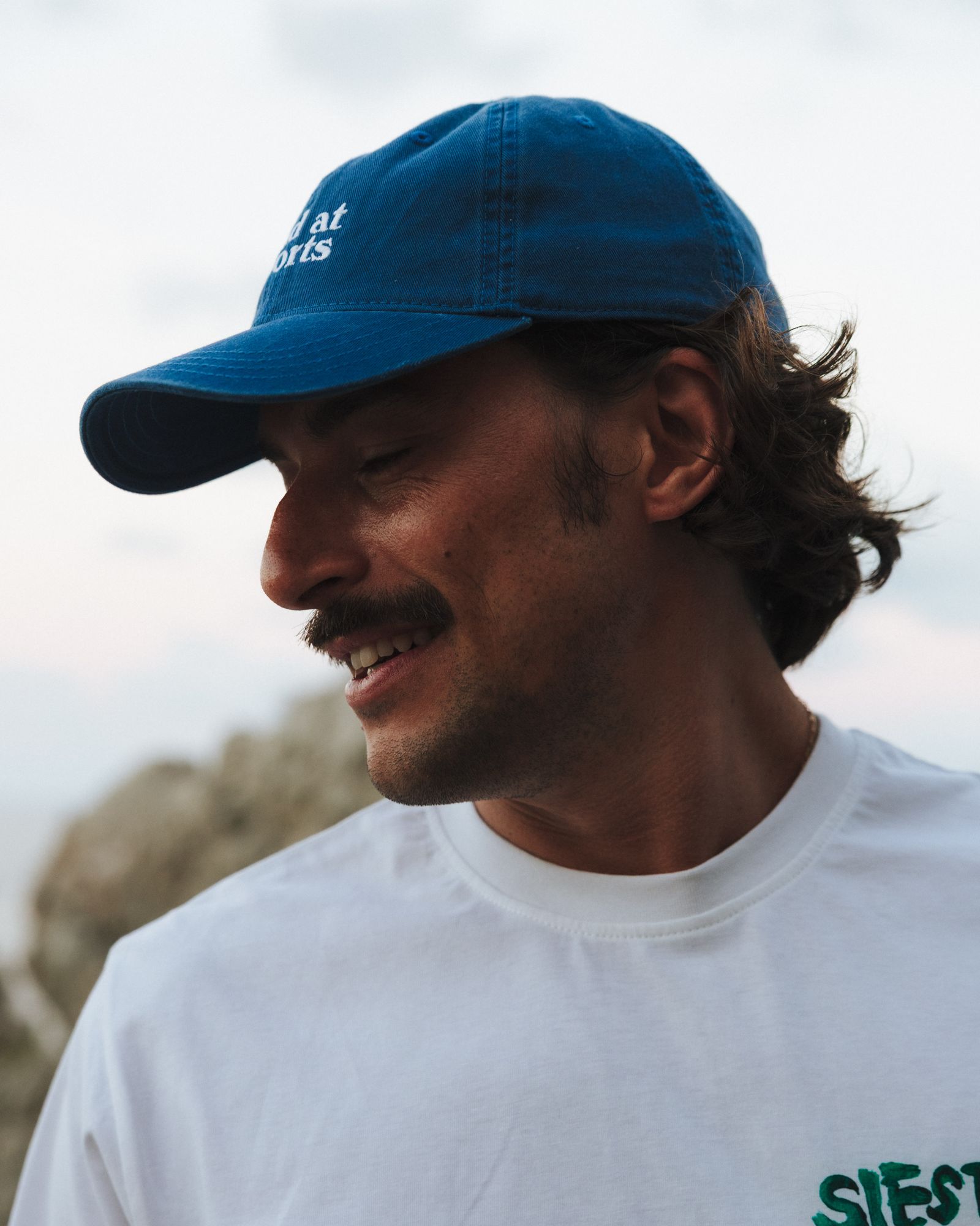 Man in a blue cap and white shirt smiles outdoors, with short curly hair and a mustache, against a blurred natural background.