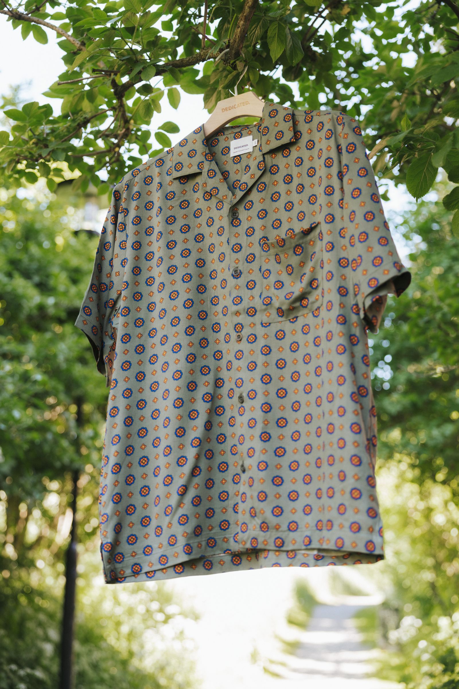 A patterned short-sleeve shirt on a hanger, displayed outdoors among green leaves and bright sunlight.
