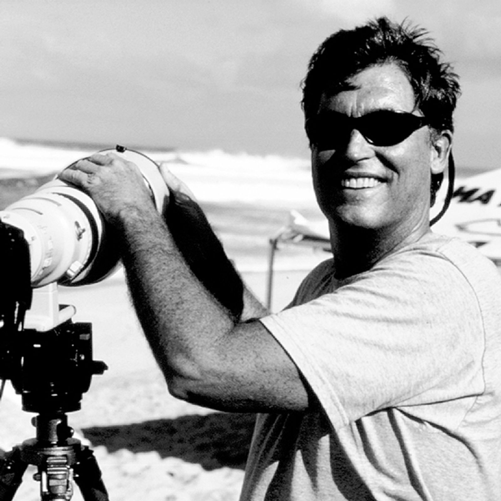 Person smiling on a beach, holding a large camera with a telephoto lens mounted on a tripod. Wearing sunglasses and a light shirt.