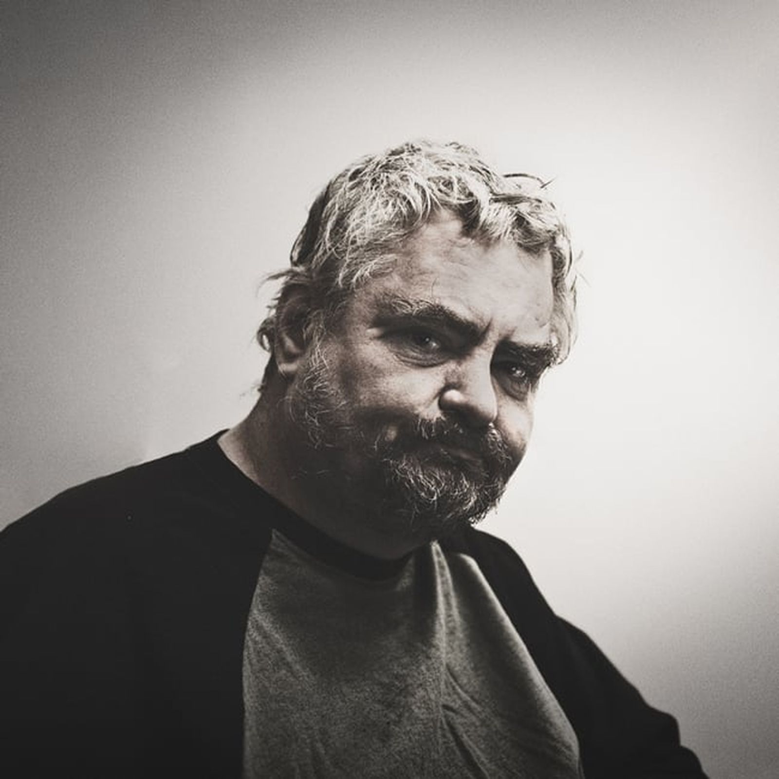 A man with a beard and tousled hair looks pensively at the camera, wearing a two-toned shirt against a plain background.