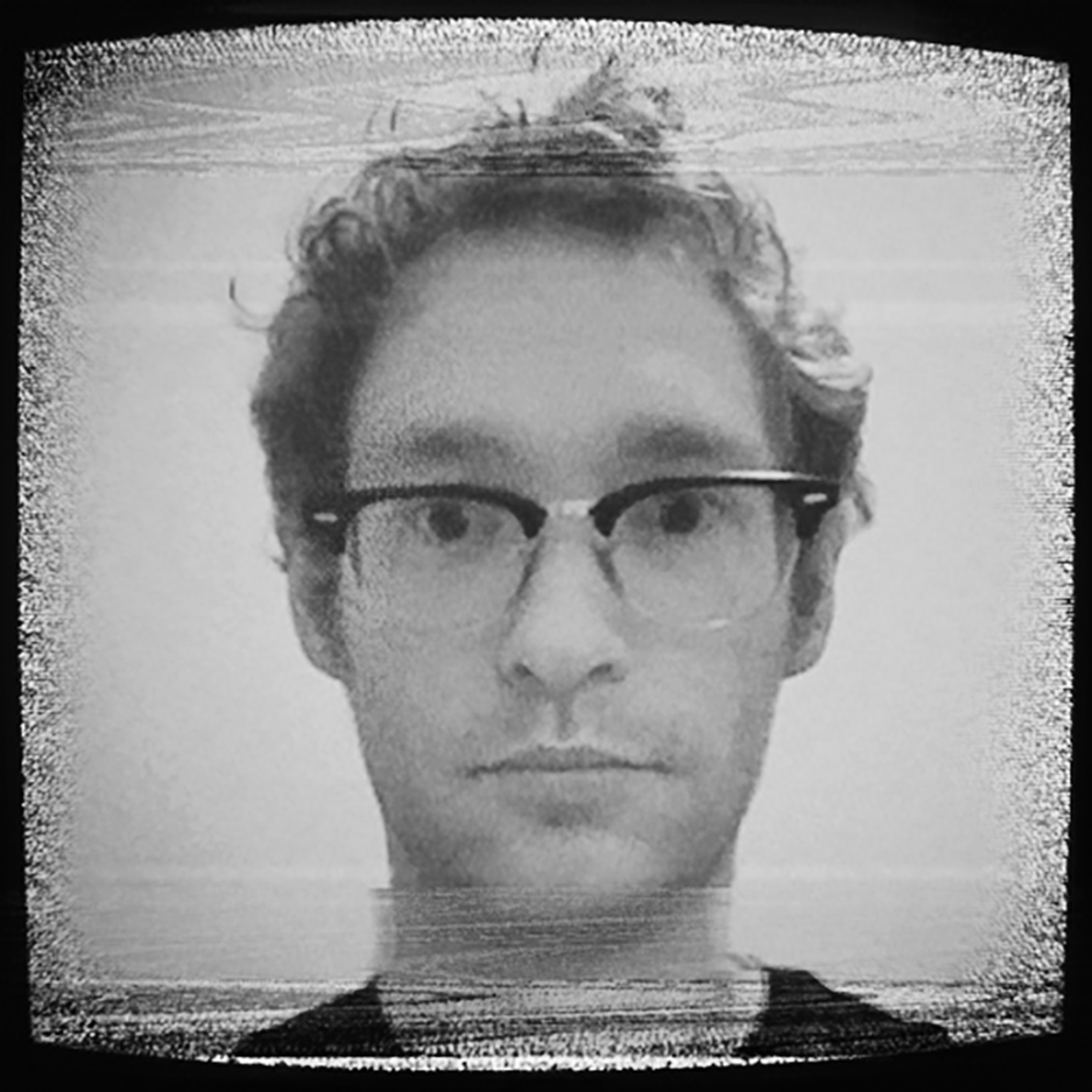 Black and white photo of a person with curly hair and glasses, looking directly at the camera with a static, grainy effect.