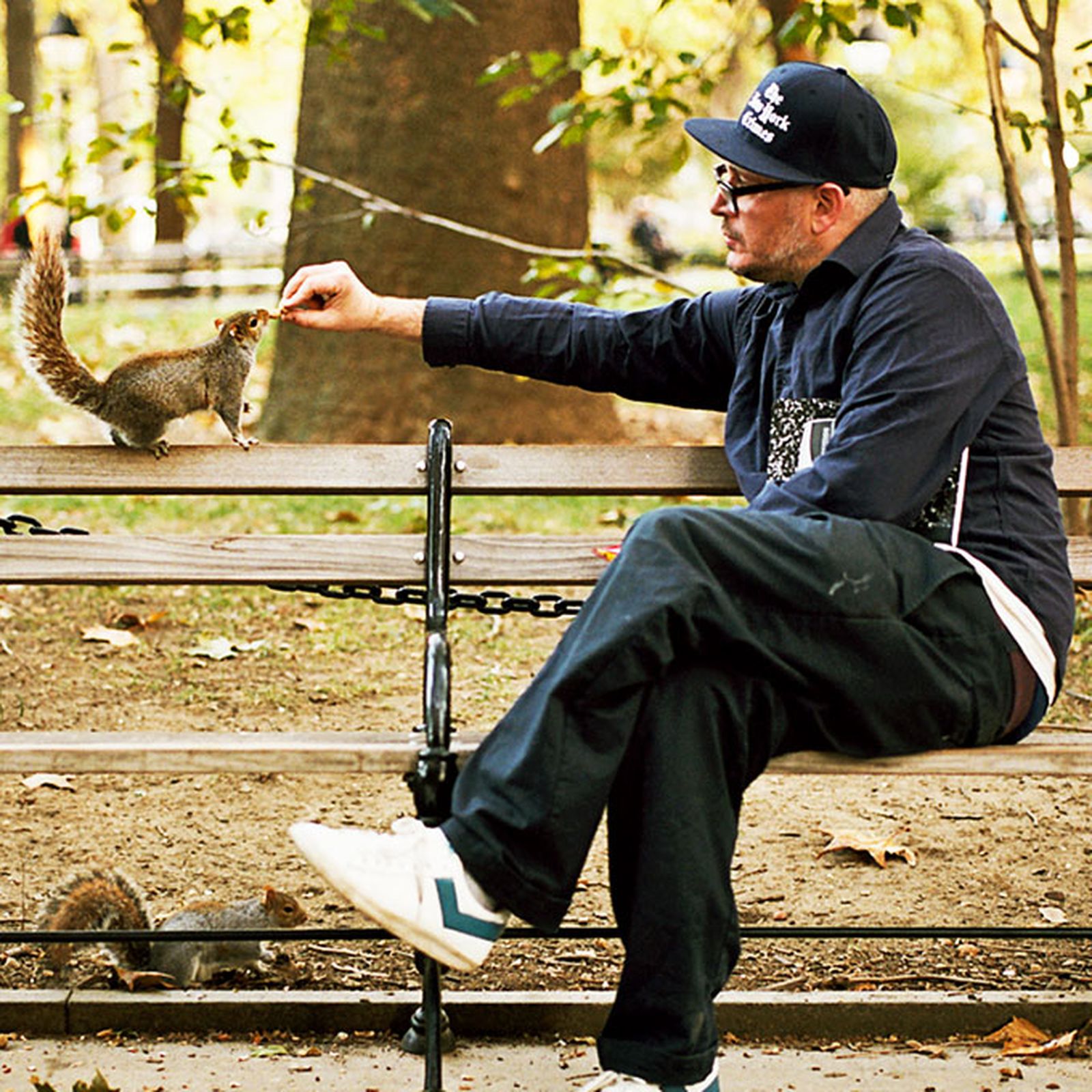 Man in casual clothing and cap feeds a squirrel on a park bench, surrounded by trees and autumn leaves.