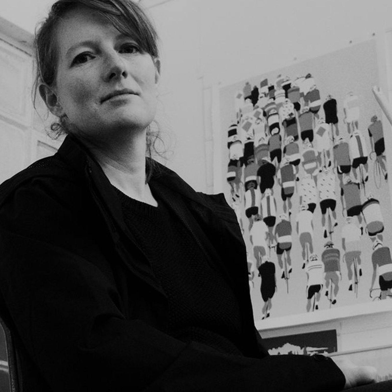 Person in a dark jacket seated indoors, with a cycling-themed artwork on the wall behind them. Black and white photo.