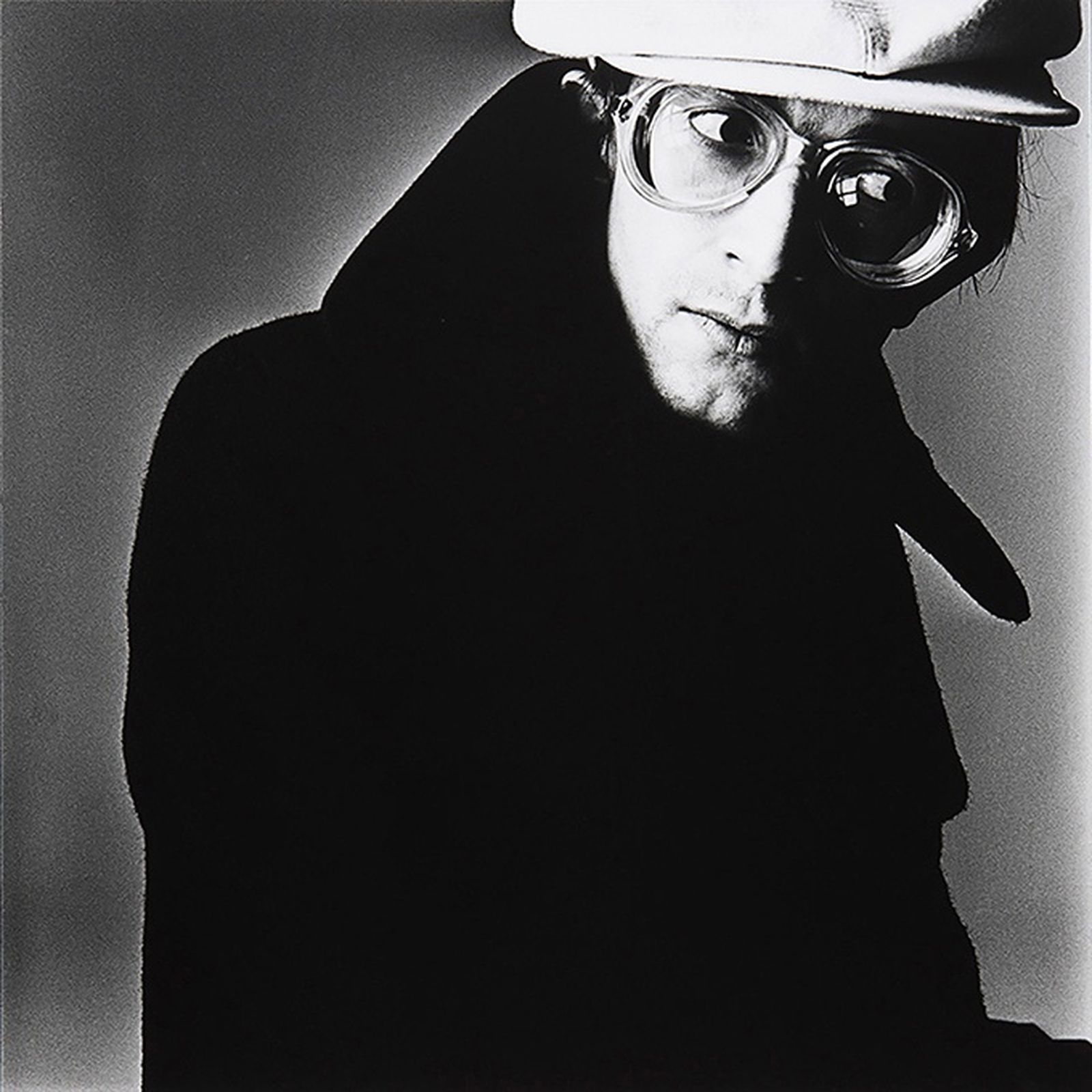 Person in a dark coat and large glasses, wearing a white hat, looking sideways against a plain background. Black and white photo.