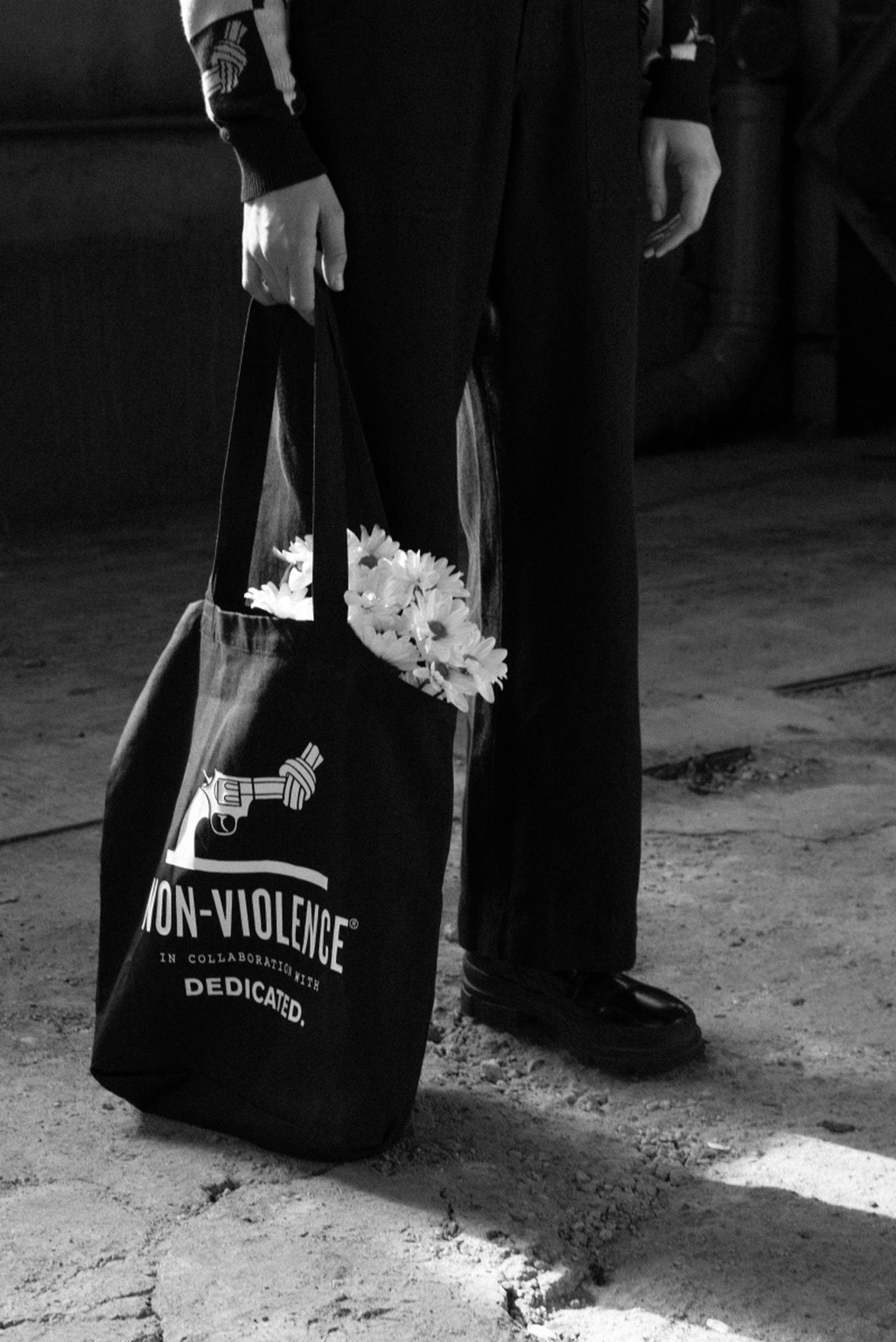 Person holding a tote bag with flowers, standing on a textured surface. The bag features a design of a knotted gun and text promoting non-violence.