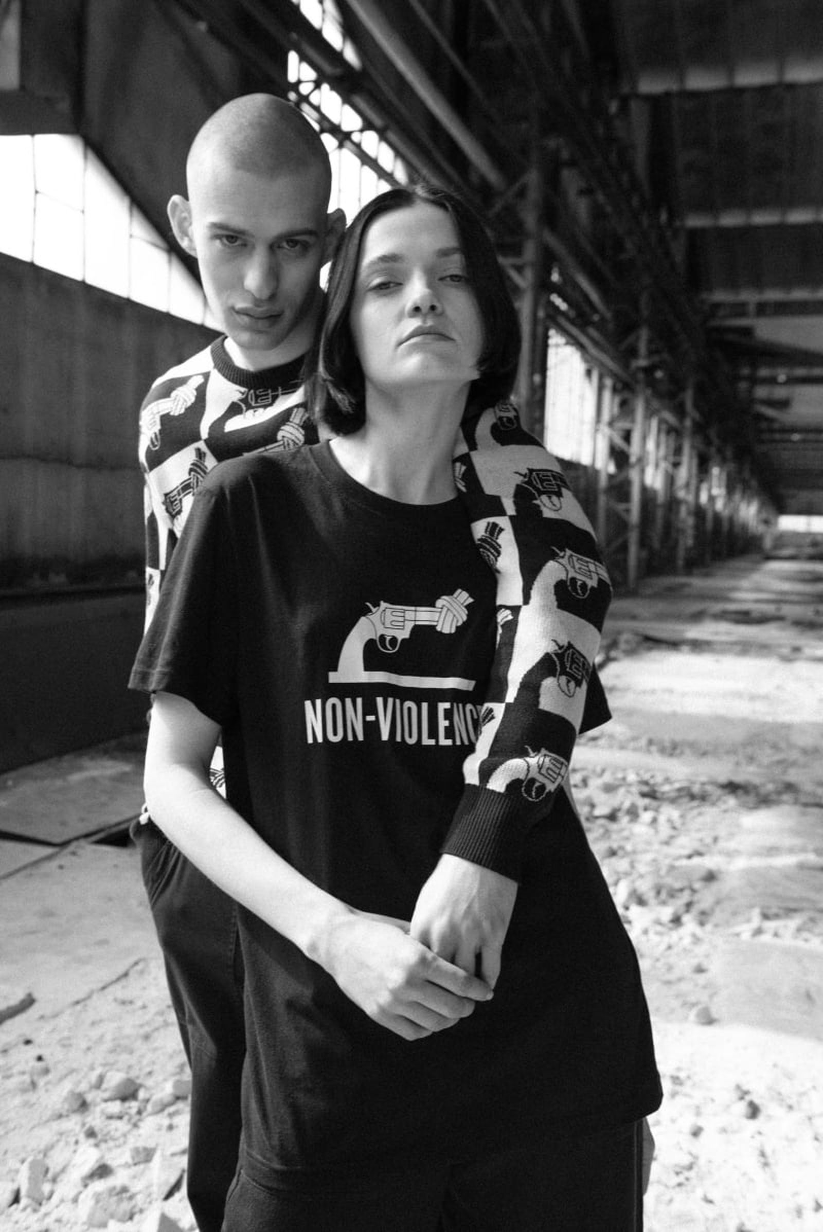 Two people stand in an industrial setting, wearing matching outfits with a non-violence symbol. One embraces the other from behind. Black and white photo.