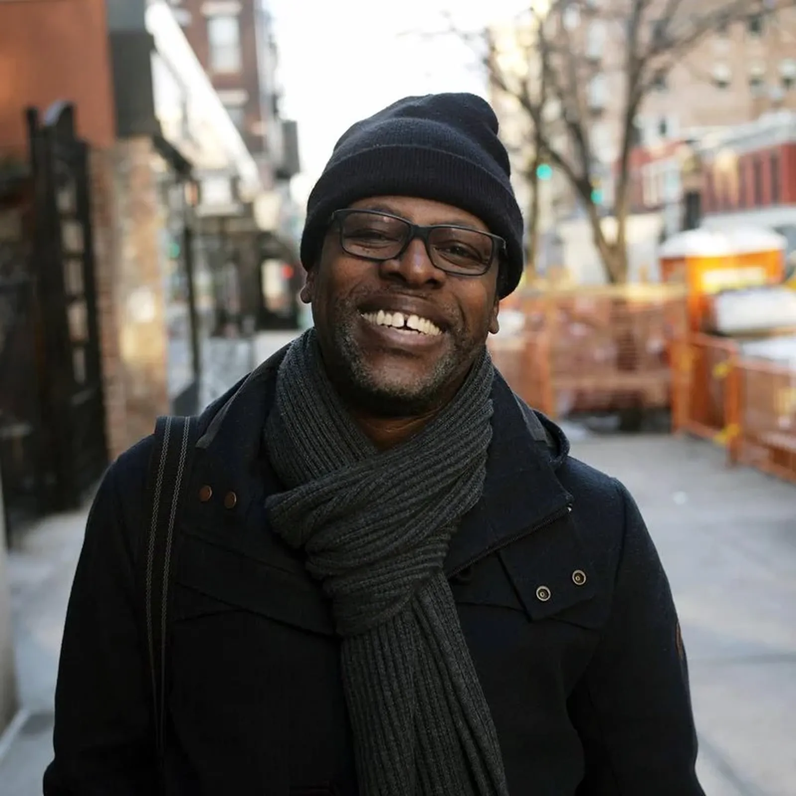 A smiling man in a black coat, scarf, and beanie stands on a city street with blurred buildings and construction barriers in the background.