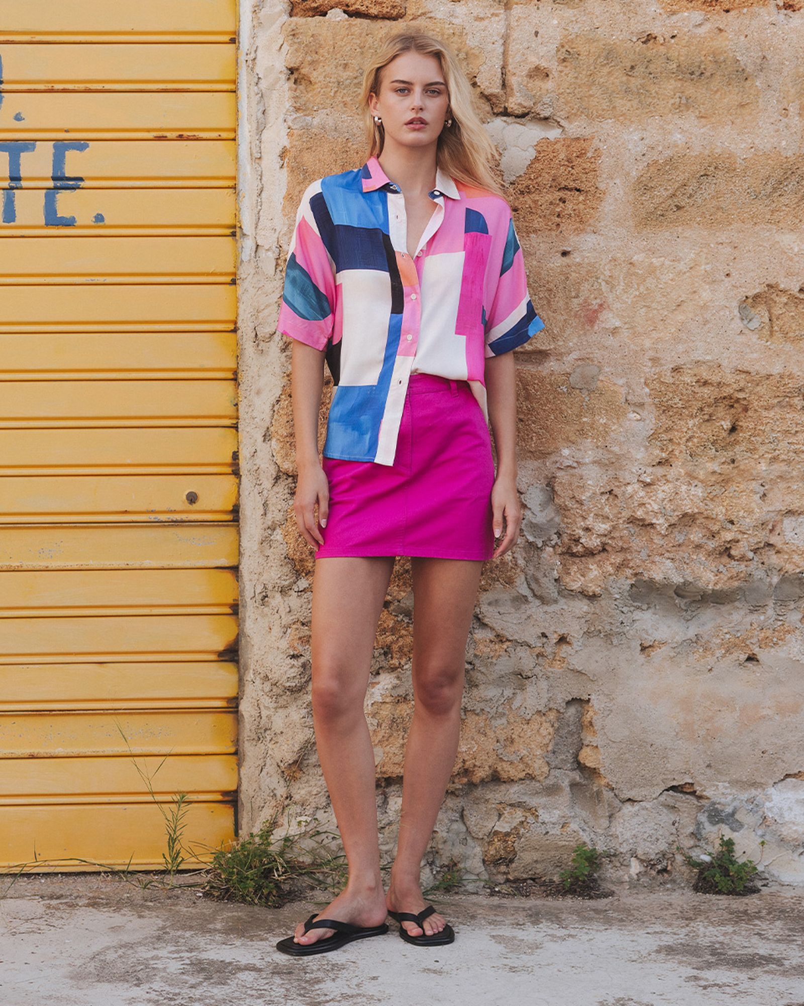 A person in a colorful shirt and magenta skirt stands against a textured stone wall and yellow door, wearing black flip-flops.
