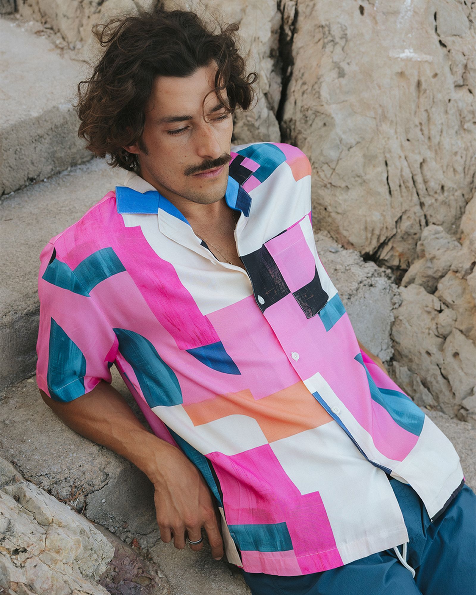 Man with curly hair and mustache wearing a colorful geometric-patterned shirt sits on stone steps, eyes closed, near rocky background.