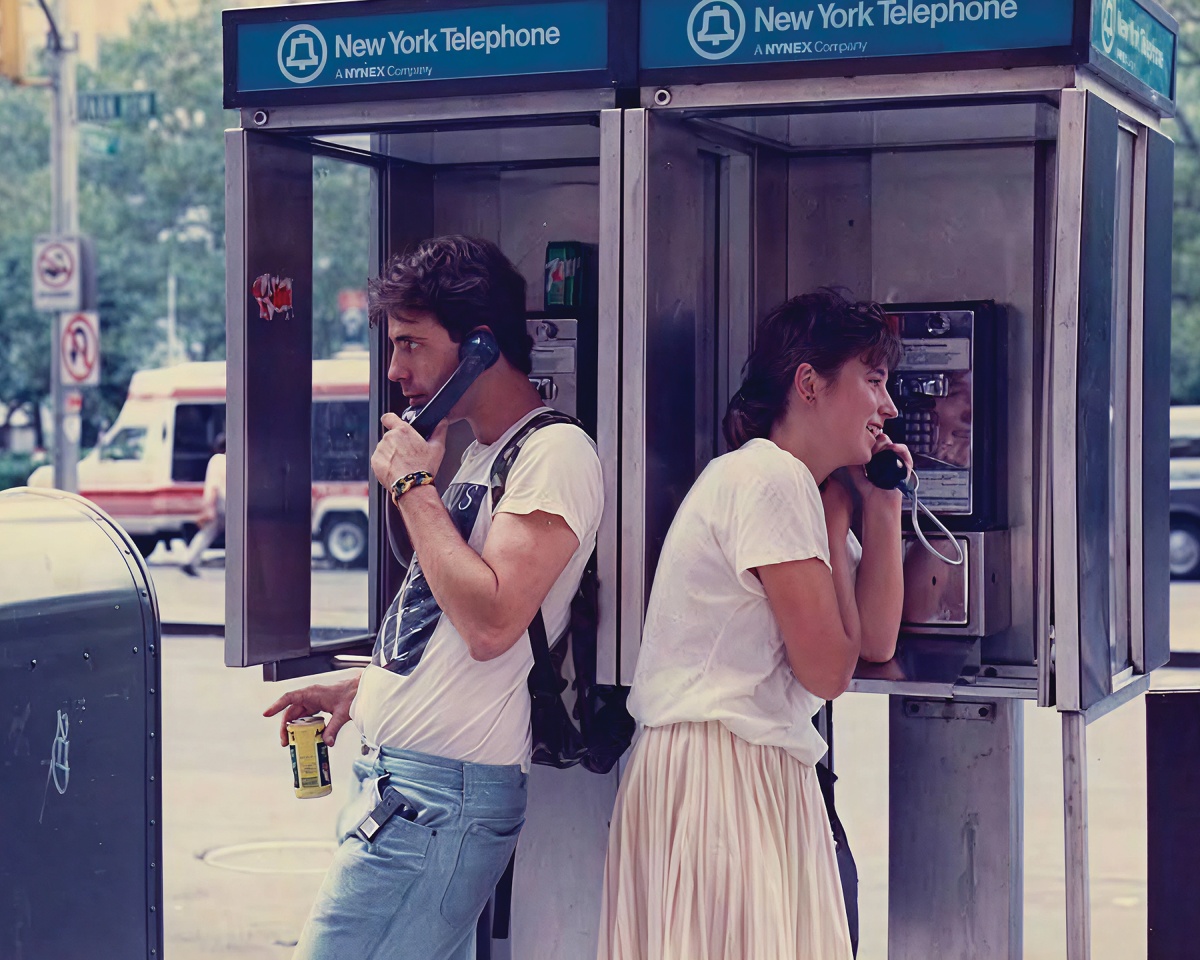 Two people using adjacent payphones labeled "New York Telephone" on a city street. The man holds a drink can, and the woman leans against the booth.