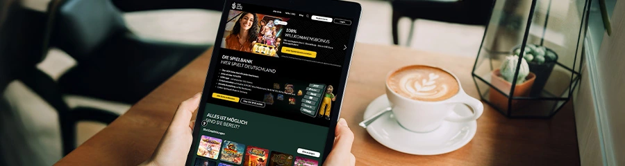 Person hält Tablet mit Casino-Website und Willkommensbonus-Banner; daneben steht eine Cappuccino-Tasse in einem Café