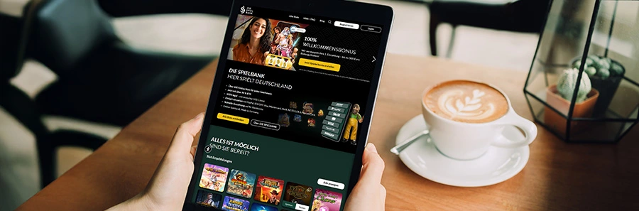 Tablet zeigt Startseite von DIE SPIELBANK mit Willkommensbonus, daneben steht eine Cappuccino-Tasse im Café.