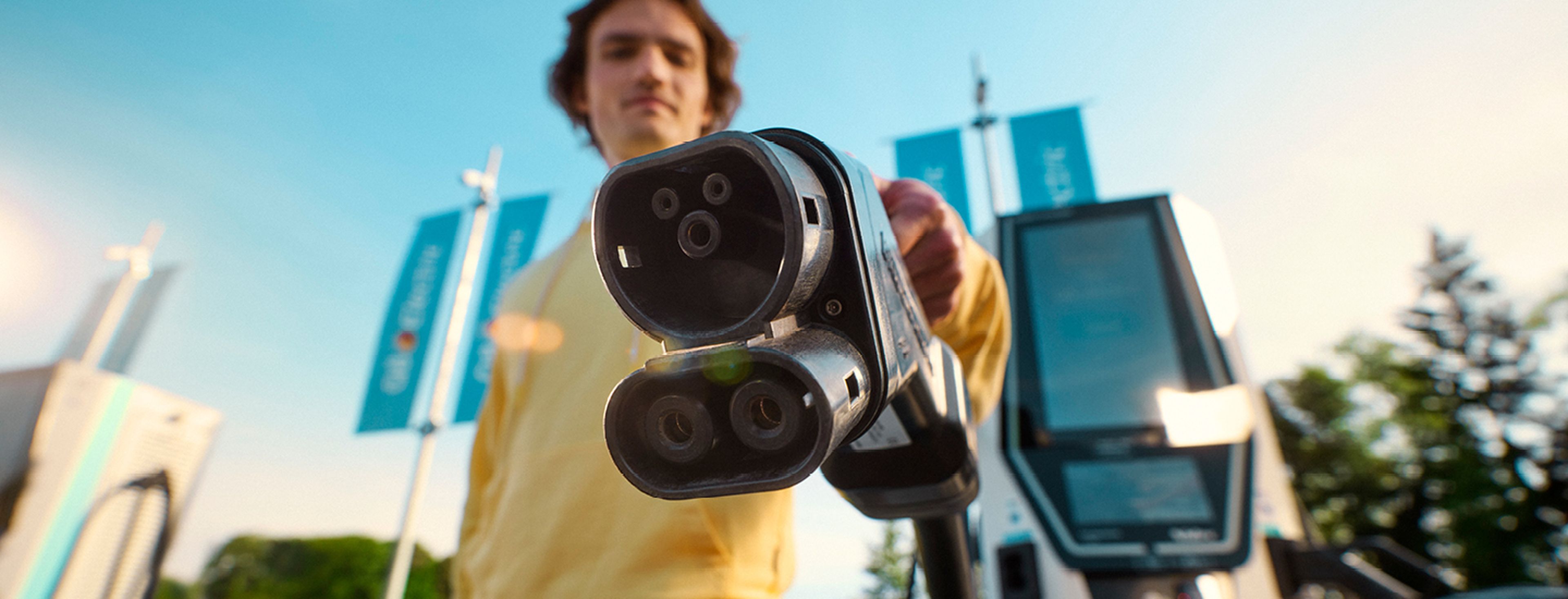 Een persoon in een gele trui houdt een EV-laadstekker naar de camera bij een Q8 electric-laadstation.
