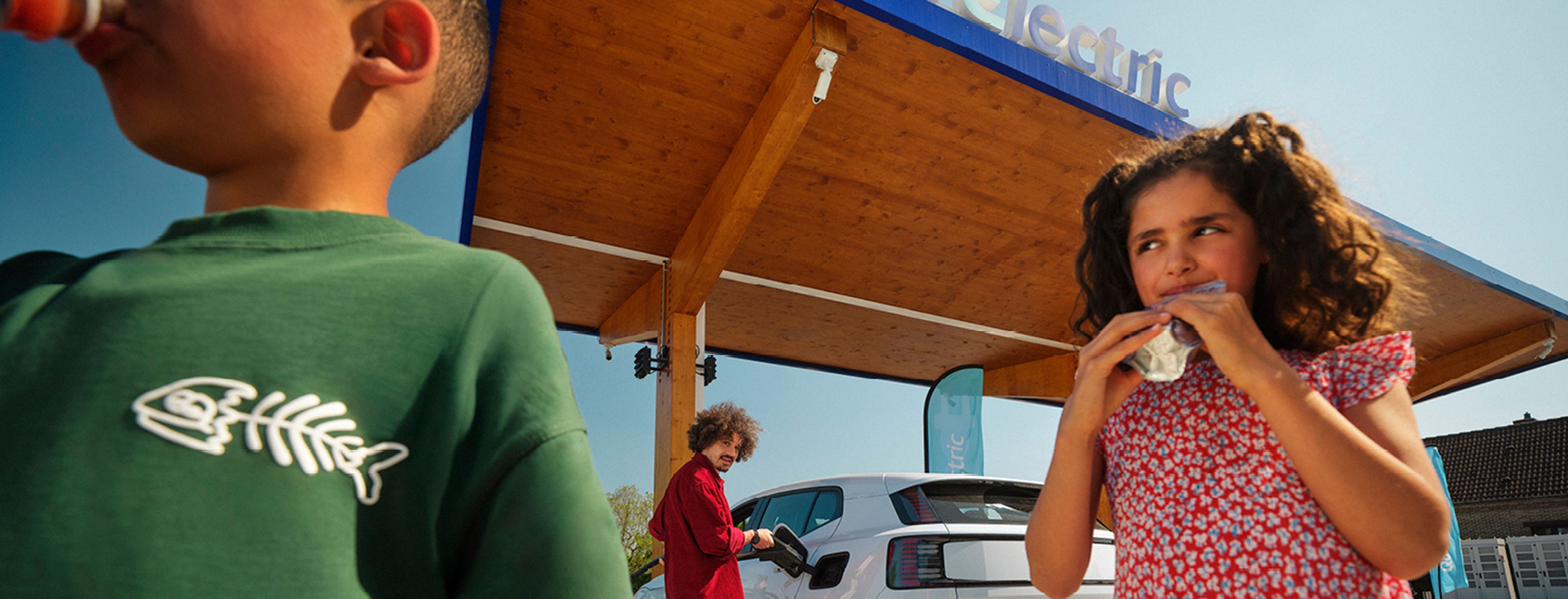 Kinderen drinken terwijl een man een elektrische auto laadt bij een Q8 Electric station.
