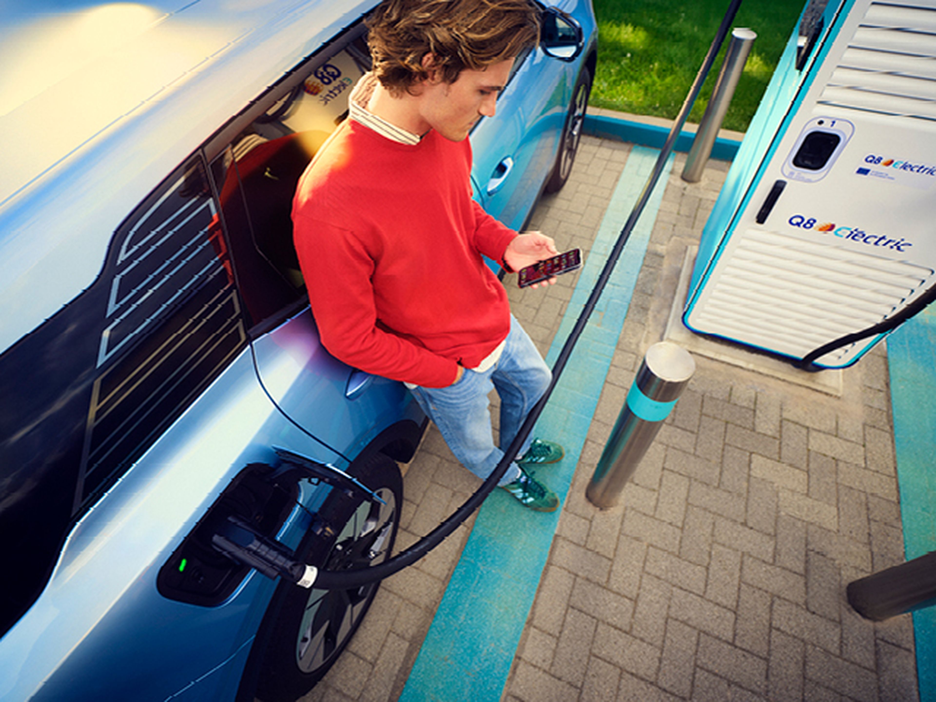 Een persoon ontspant zich in een geparkeerde auto bij een Q8 electric laadstation, met de voeten uit het raam en een koffiebeker in de hand. Op de achtergrond zijn Q8 electric vlaggen en een snellader zichtbaar.